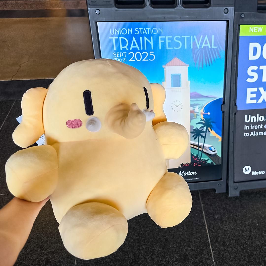 Photo of the Creature Mastodon stuffed toy held in front of a “Union Station Train Festival” sign under bright, natural lighting indoors during the day. 
