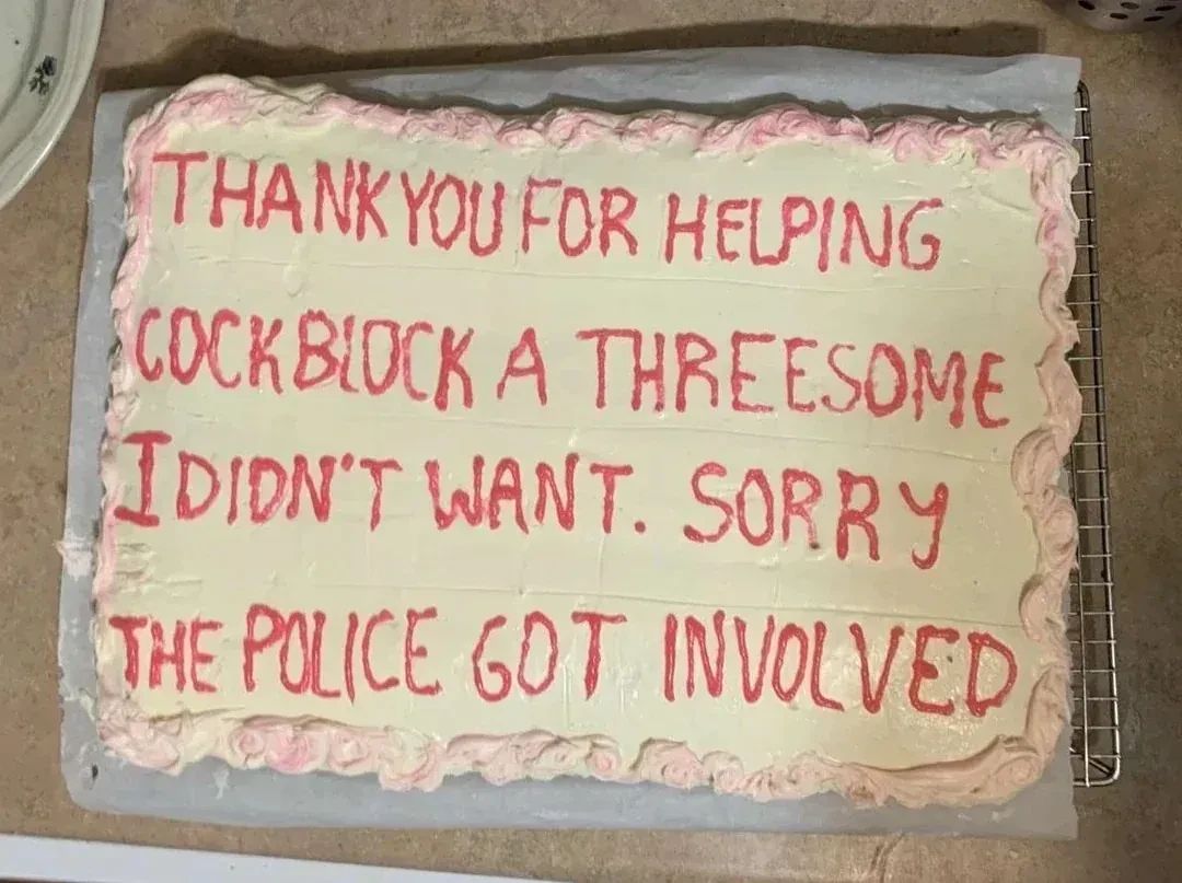 The image shows a rectangular cake with white frosting and red lettering. The cake is sitting on a metal cooling rack, which is placed on a surface that appears to be a countertop. The cake is decorated with pink frosting along the edges in a wavy design. The red lettering on the cake reads, "THANK YOU FOR HELPING COCKBLOCK A THREESOME I DIDN'T WANT. SORRY THE POLICE GOT INVOLVED." The lettering is somewhat uneven, suggesting it was written by hand. The composition is a close-up shot, focusing on the cake and its message. The color palette consists primarily of white, pink, and red, with the background providing a muted brown tone. There is a plate with a blue floral pattern visible in the top left corner.