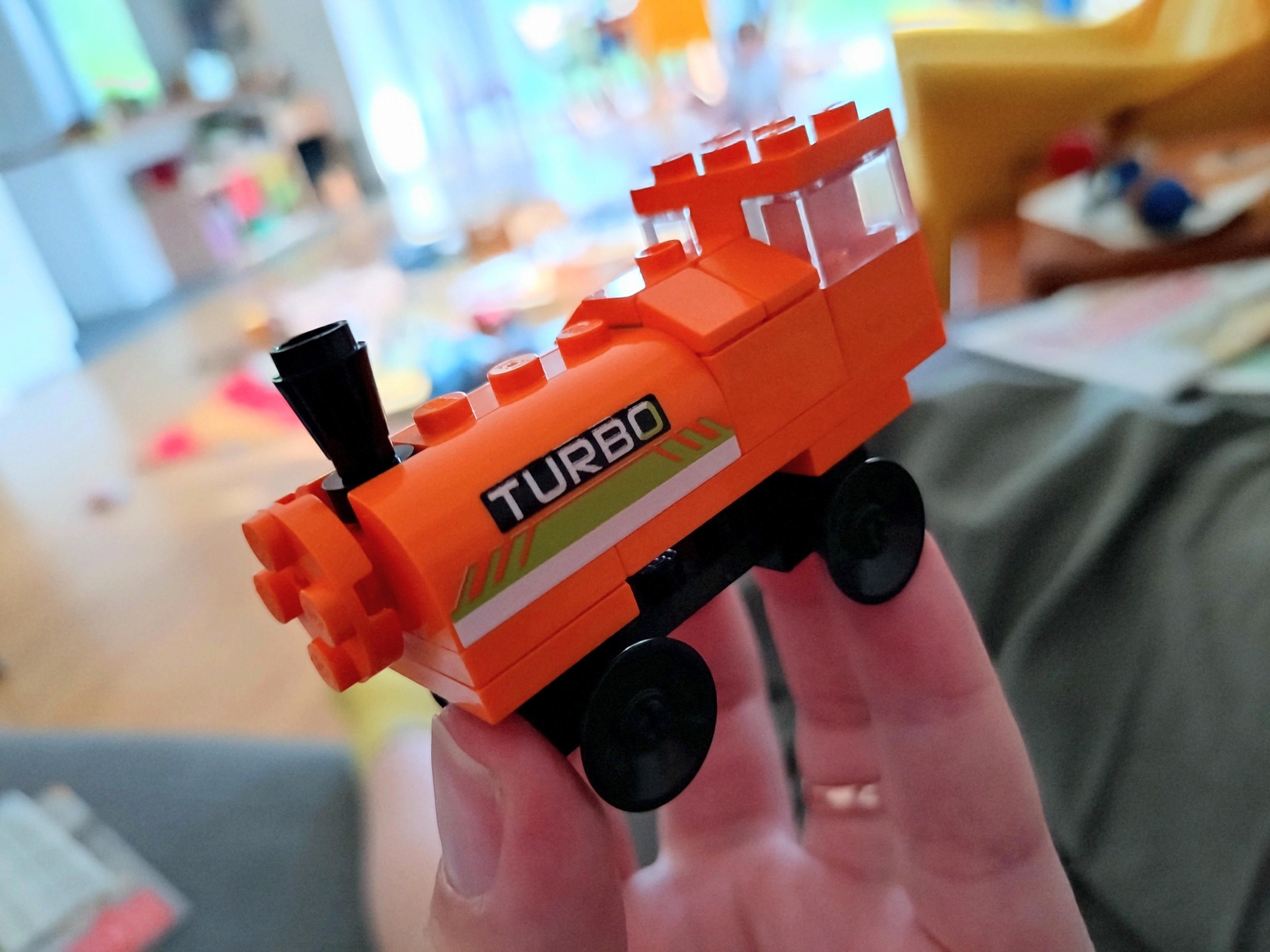 An orange steam engine built with Lego. It is 3 studs wide, 8-ish studs long, in saddle-tank style with a larger section where the firebox is. Amusingly the curved part of the water tank has "TURBO" printed on it, presumably these bricks came from a racecar set.