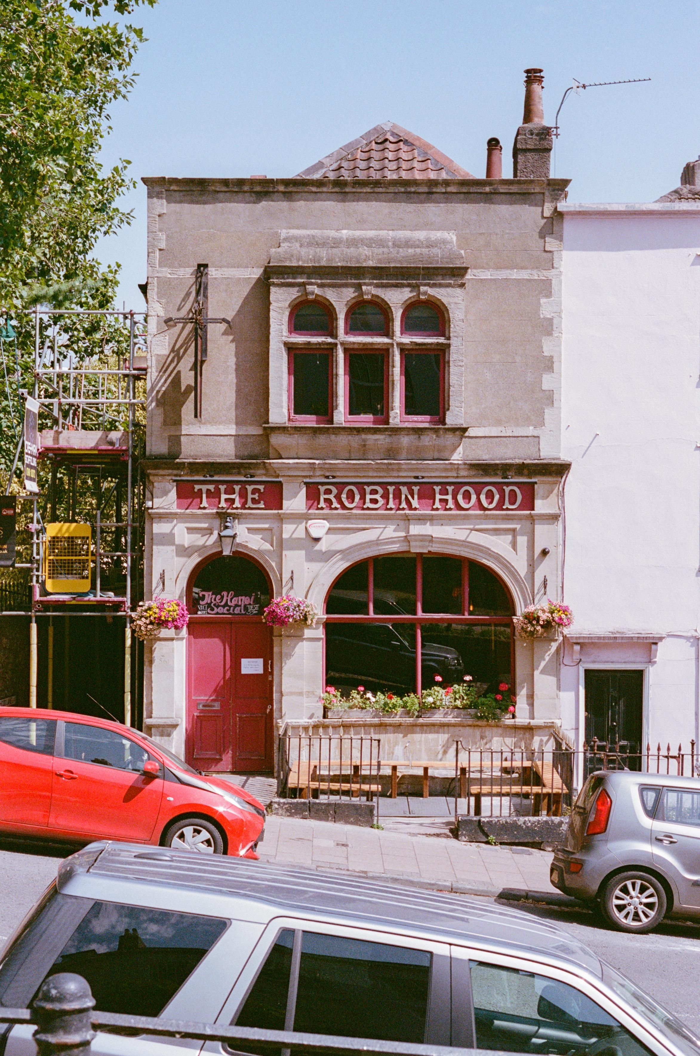 A two story building with a red door called "The Robin Hood", with flowers under a big street level window, on a steep road.