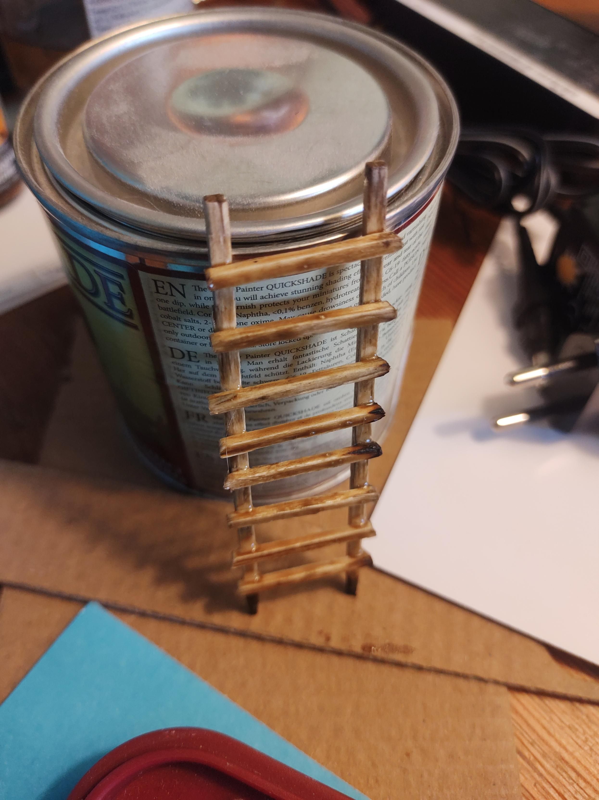 A little wooden ladder made out of match sticks, propped up against a small pot of enamel paint. The ladder looks shiny and sticky, with a light brown tone.