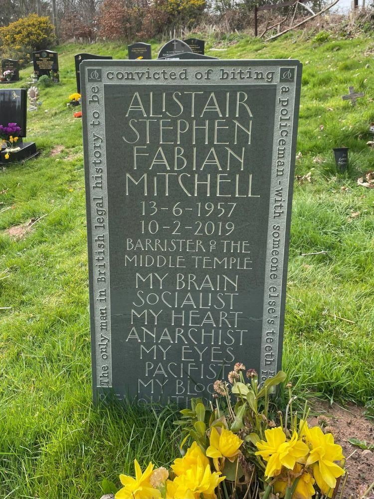 Gravestone inscribed: Alistair Stephen Fabian Mitchell. 13-6-1957, 10-2-2019. Barrister of the Middle Temple. My brain, socialist. My heart, anarchist. My eyes, pacifist. My blood, revolutionary. 

Around the edge, the legend: The only man in British legal history to be convicted of biting a policeman with someone else’s teeth.