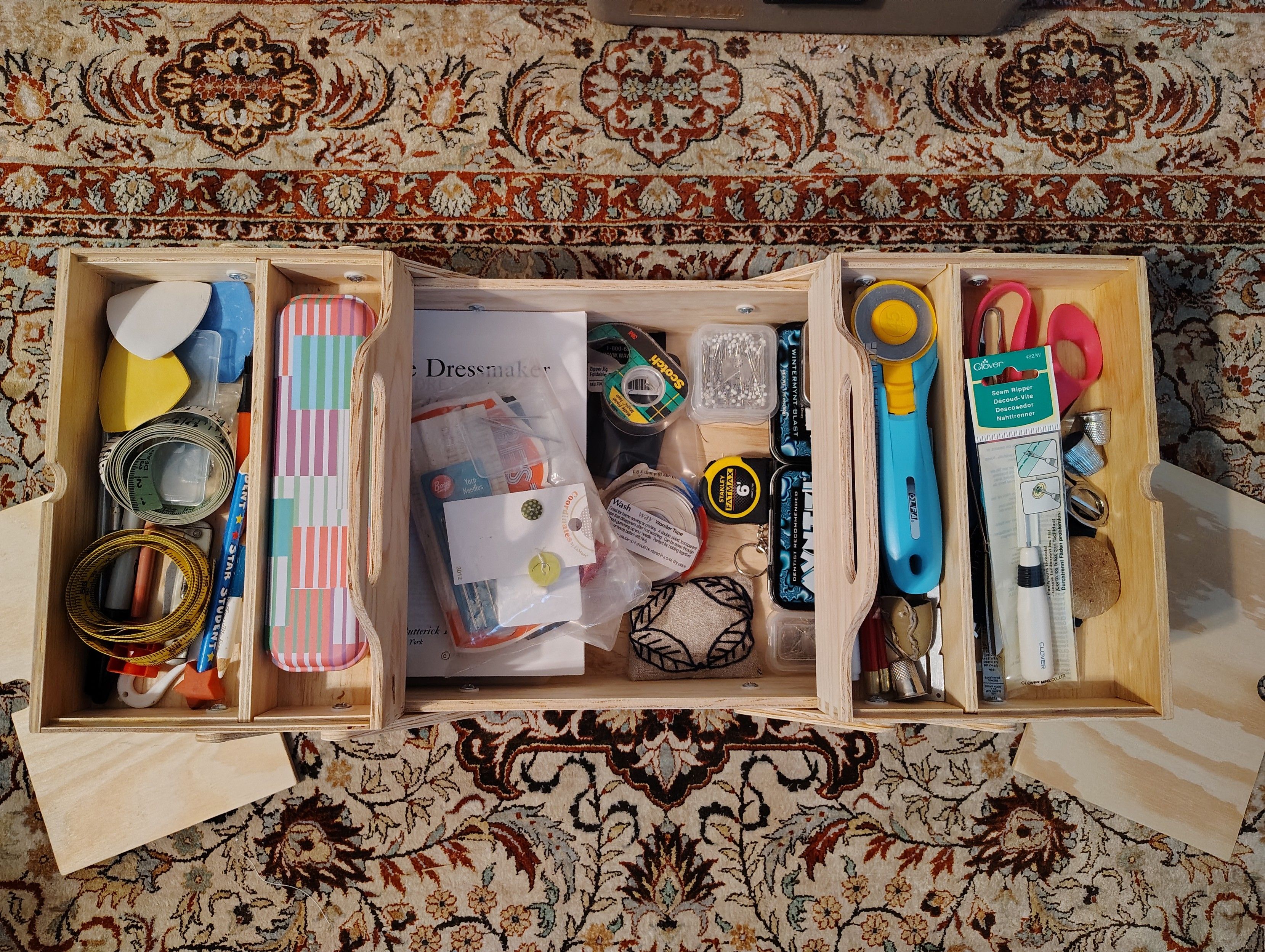 A large wooden cantilevered tool box filled with sewing notions (including scissors, measuring tapes, marking chalk and rotary cutter).