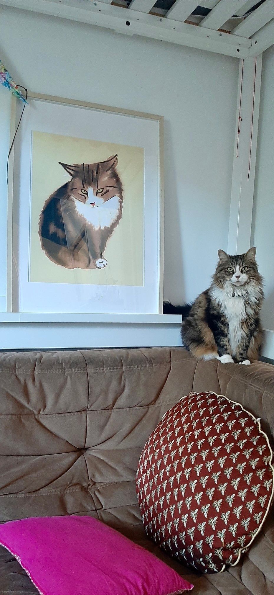 Chatte assise sur le dossier d’un canapé marron devant un portrait d’elle plus que grandeur nature que j’ai dessiné. Elle regarde droit dans l’objectif.