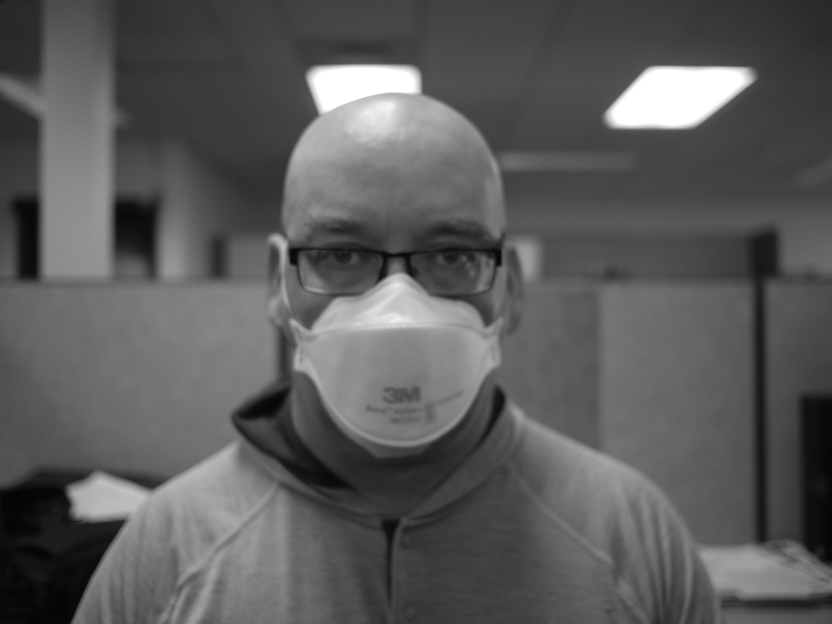 A bald man is staring at the camera in a black and white picture. His eyes are behind squared off glasses and his lower face is obscured by a 3M Aura Mask. Above his glasses a pair of lush, dense eye brows underline the smooth shiny dome of his head. Behind him can be seen cubicle walls, support columns and fluorescent lights shining out of a drop ceiling made of acoustic tile.
He is wearing a one piece shirt with an integrated hood folded down around his neck.