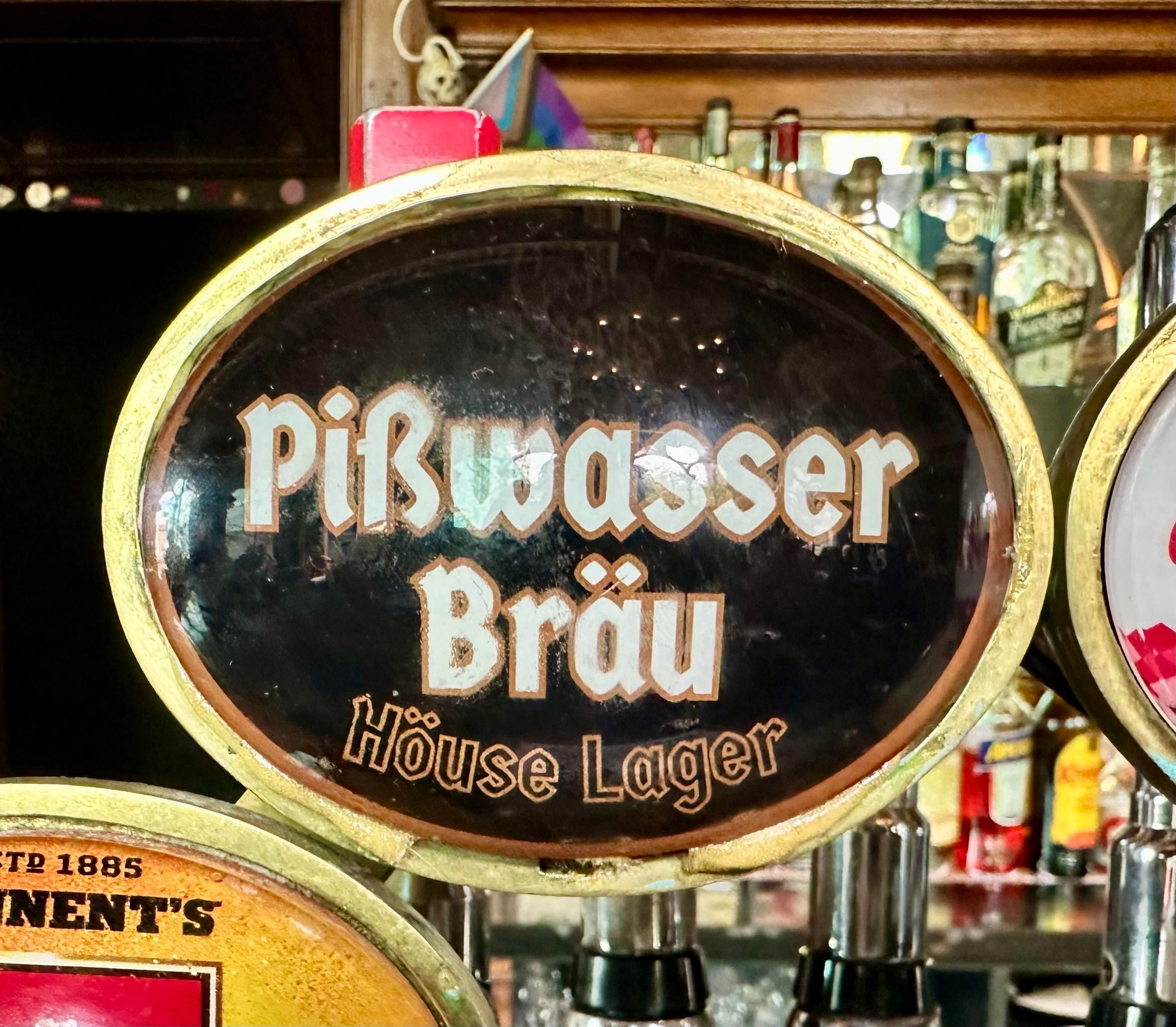 Photo of a keg beer tap with a black label and white blackletter words saying “Pißwasser Bräu House Lager”, with a heavy metal umlaut on the o of “House”.