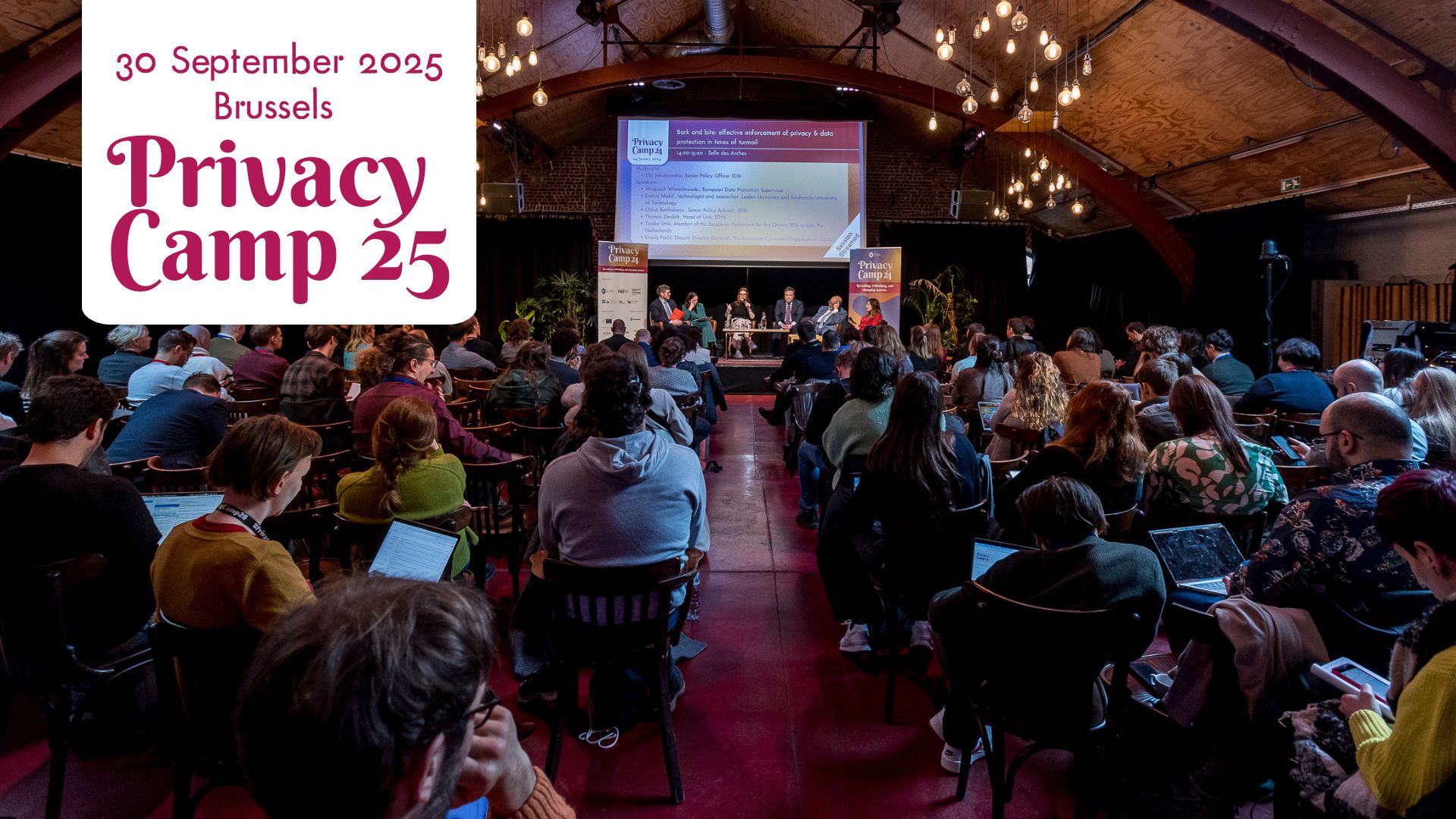 The photo shows a panel in progress at Privacy Camp in 2024. In the centre, there are six speakers on the stage and we can see a full room of audience from behind engaged in the session. The room is rustic and woody with bulbs hanging from the ceiling. In the top left corner, there's a banner: 30 September 2025, Brussels, Privacy Camp 25.