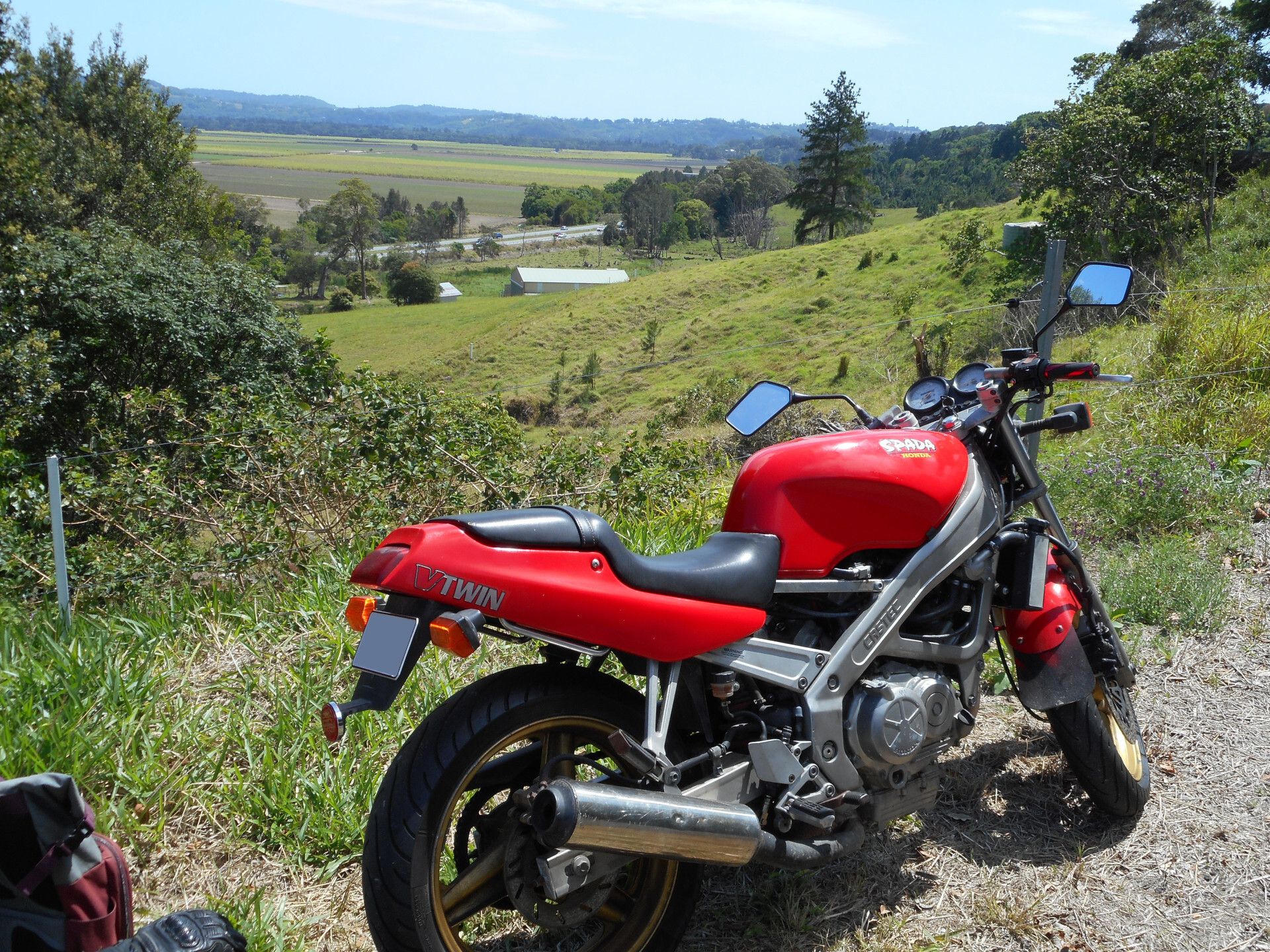 A red Honda VT250 Spada, parked overlooking the Pacific Highway from a dirt road above it. It has no fairings, modern style mirrors, a "SPADA" decal on the tank, and "V-TWIN" on the tail.