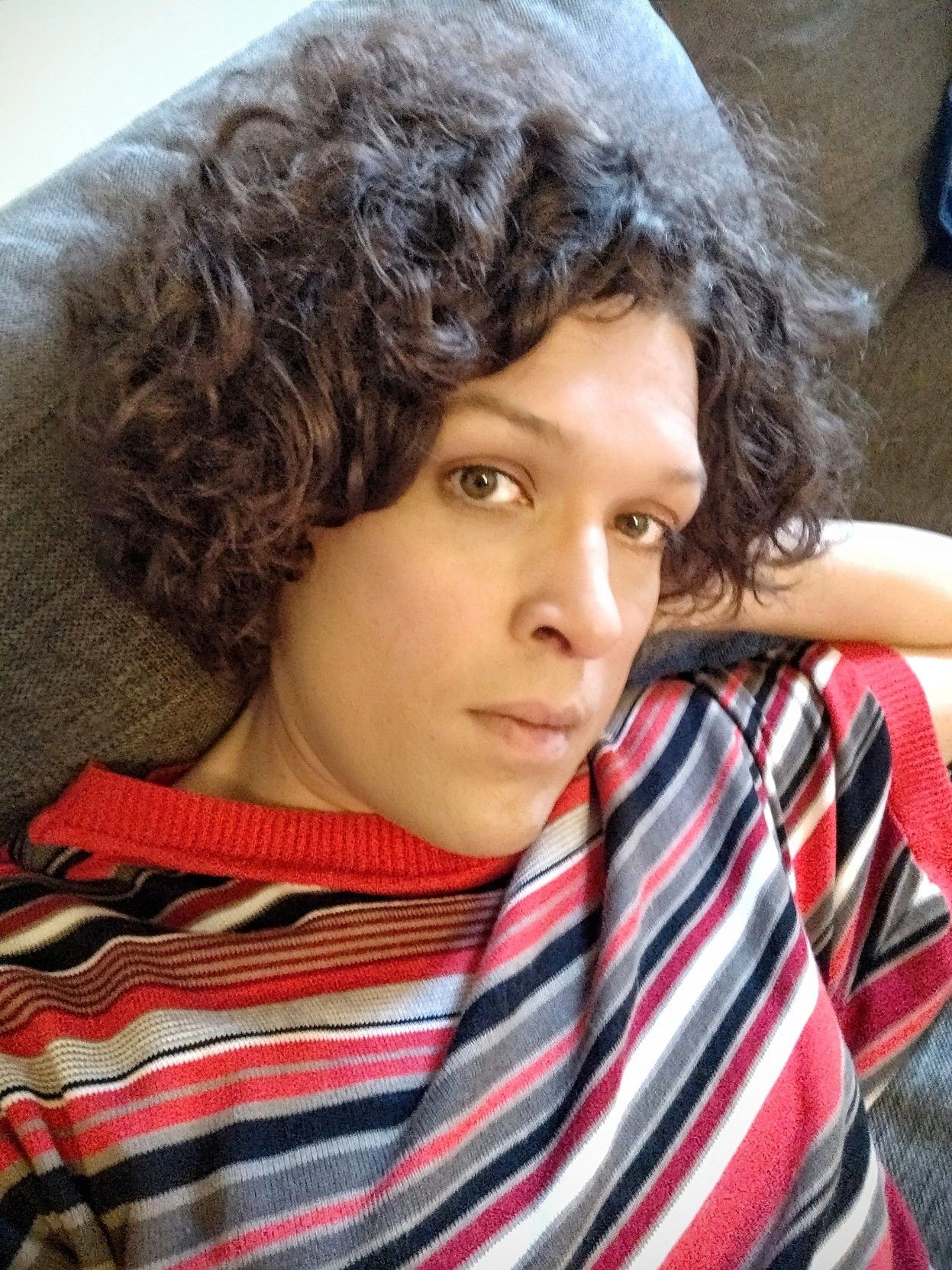 Me leaning back and almost lying down in my sofa with my left hand behind my back, looking thoughtful and a bit tired, wearing one of my favourite tops, with wide and short sleeves, and horisontal stripes of different width, in red, white black and grey.