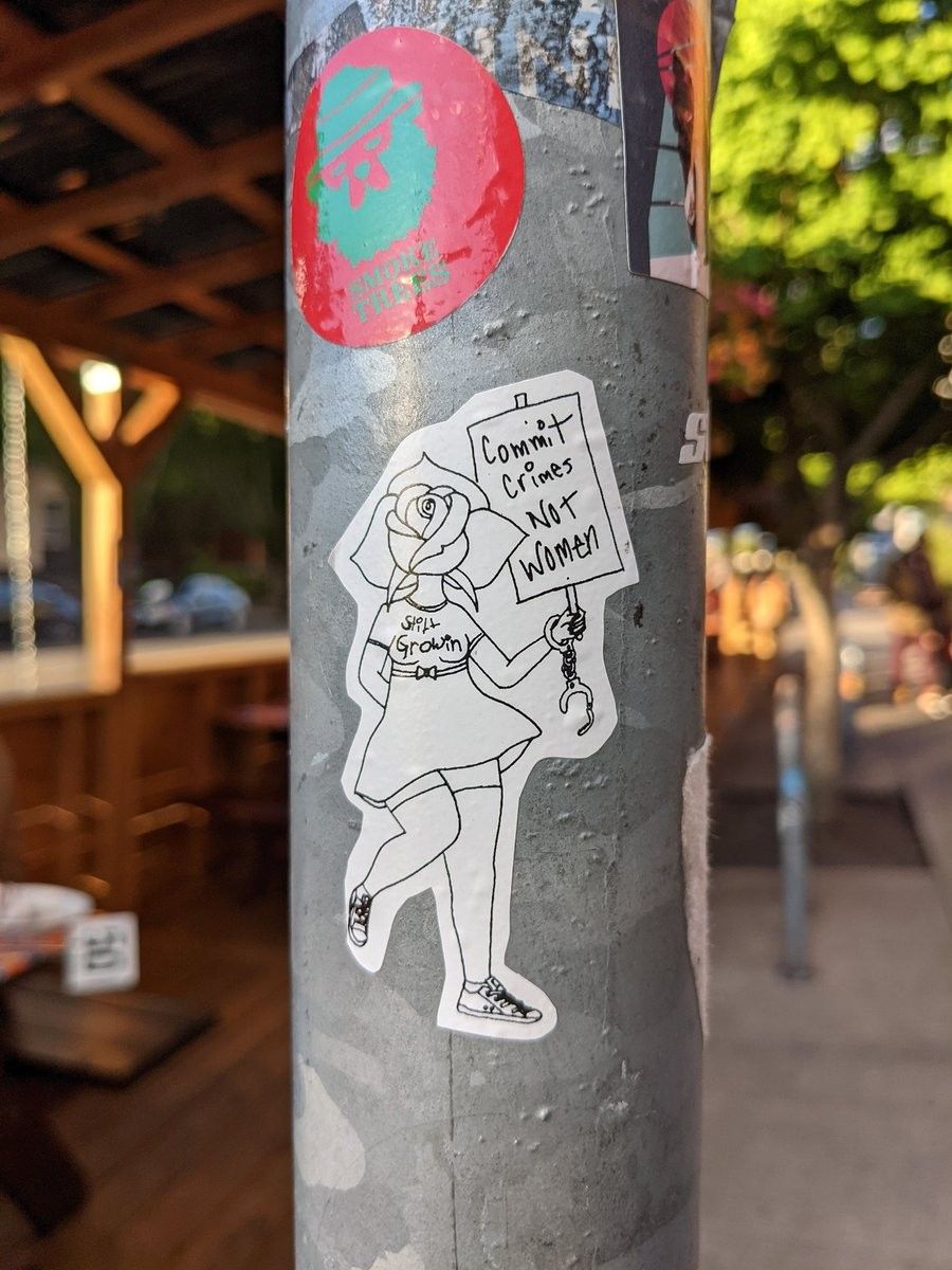 A sticker on a metal pole showing a girl with a rose for a head wearing a dress. She carries a sign that says, "Commit crimes, not women."