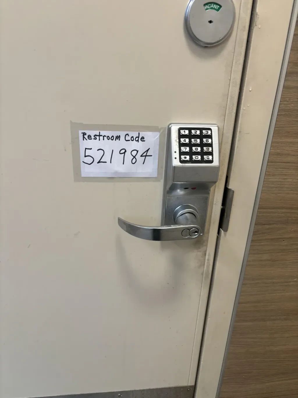 The image showcases a portion of a restroom door. The door is painted in a light, off-white color and features a combination lock and a handle.

A handwritten sign, taped to the door, indicates "Restroom Code" followed by the number "521984". The sign is made of plain white paper and the text is written in black marker. The combination lock is a metallic gray color and has a keypad with numbers 1 through 9, an asterisk, and a zero. Above the lock is a circular indicator with the word "VACANT" visible, suggesting the restroom is currently unoccupied.

The door handle is made of a brushed metal, and the edges of the door appear to have some wear and tear. The surrounding wall and floor are visible in the background, with the floor appearing to be a light brown with a wood grain pattern. The composition is a medium shot, focusing on the details of the door and its hardware. The lighting is bright and even, casting minimal shadows.