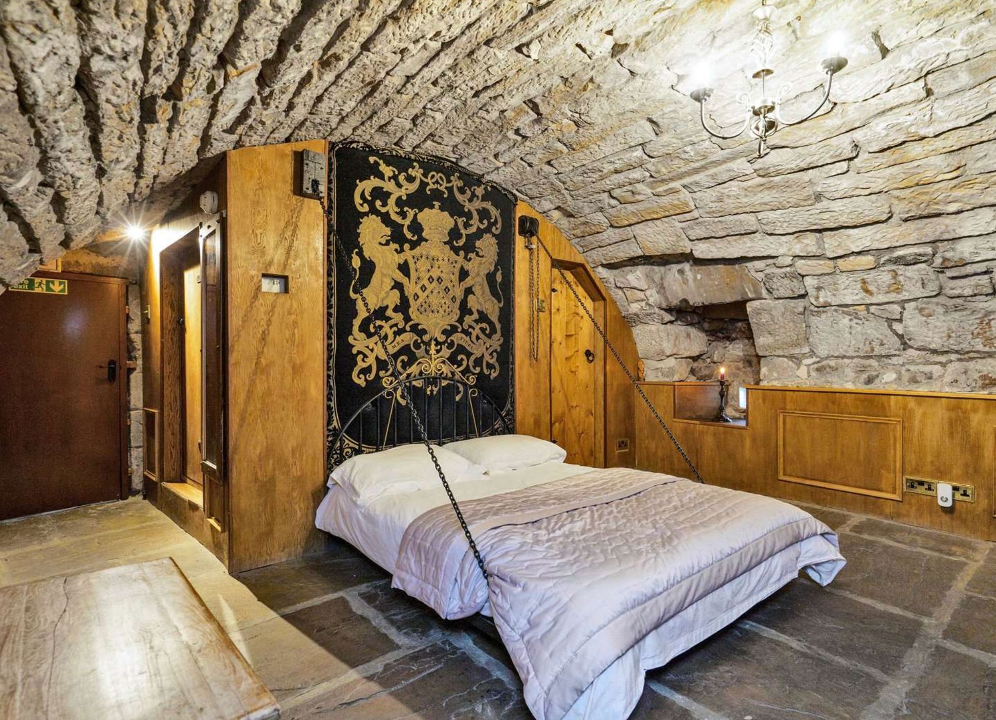 A bedroom with a curved stone ceiling and a bed that has chains attached to either side, attached to the wooden wall. The chains look like they'd be in the way if you were getting in or out of bed. 