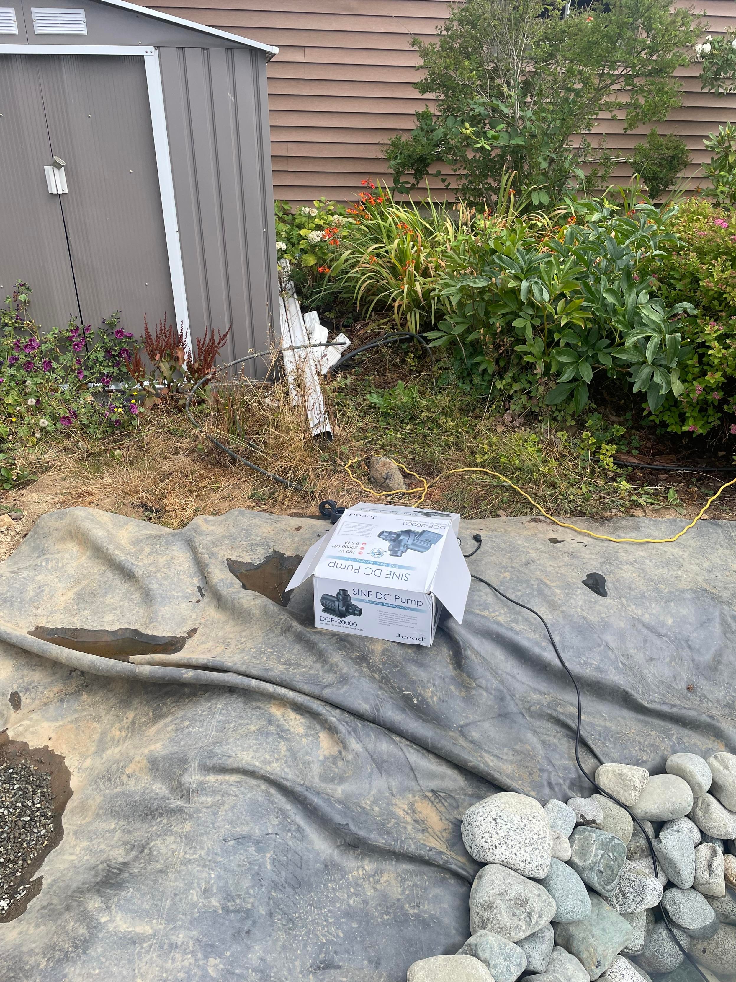 A small box with the picture of a pump sits on the corner of the pond. There is a small black wire leaning towards it. In the background is a small gray shed beside a flower garden.
