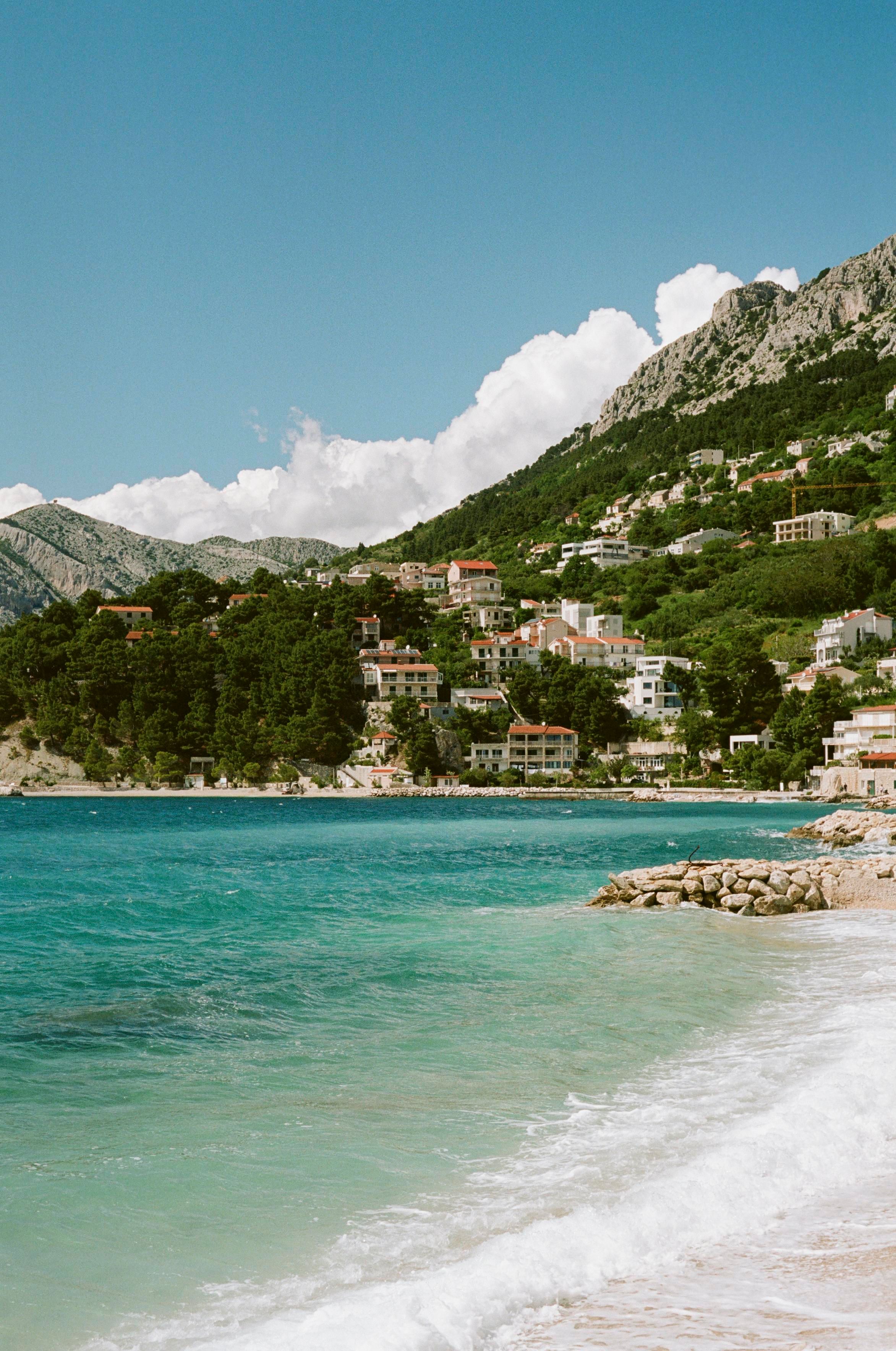 Turqoise sea, foaming as it approaches the coast. A village with red roofs, enroaching on a mountain. More mountains behind, with low set clouds barely making it over the mountains. Above, a bright blue sky.