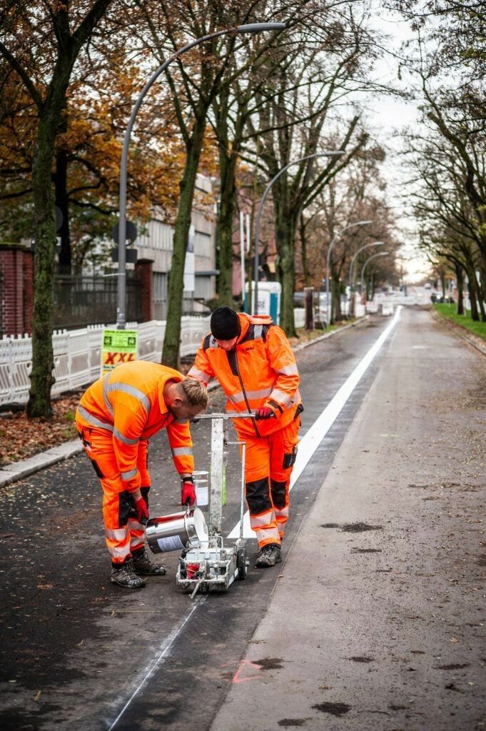 Foto: Zwei orange gekleidete Bauarbeiter bringen auf einer herbstlichen Allee einen weißen Streifen auf die Fahrbahn. 