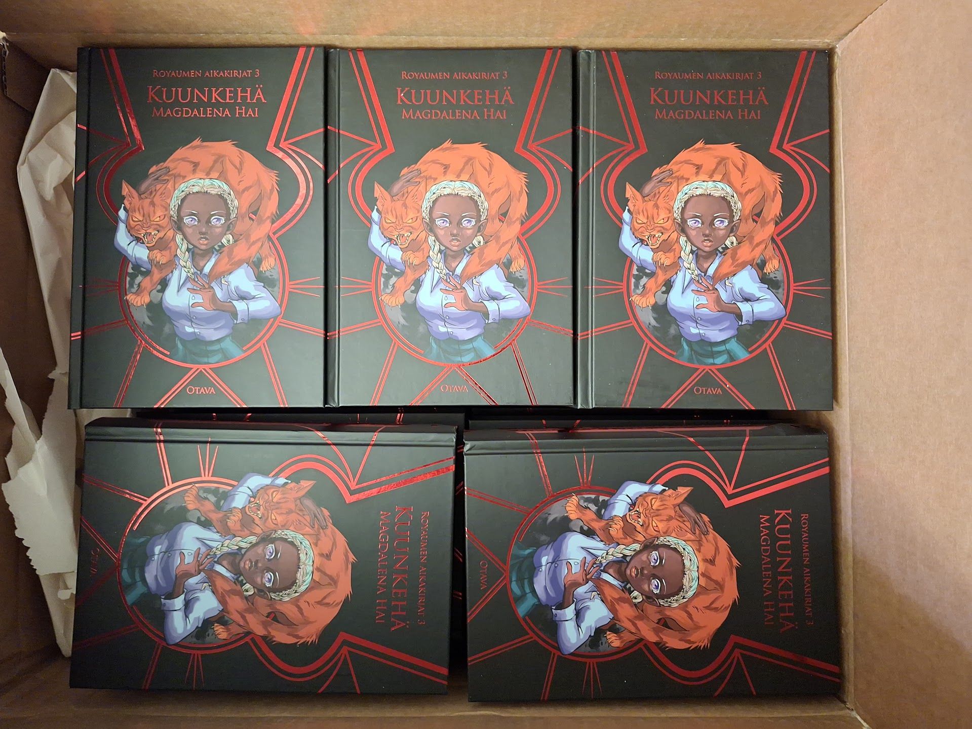 Näkymä isompaan laatikoista: viisi kirjaa, joiden kansi on musta punaisin, metallisin aksentein ja jonka keskiössä on tummaihoinen, valkotukkainen tyttö vihaisen näköinen punainen kissa harteillaan.