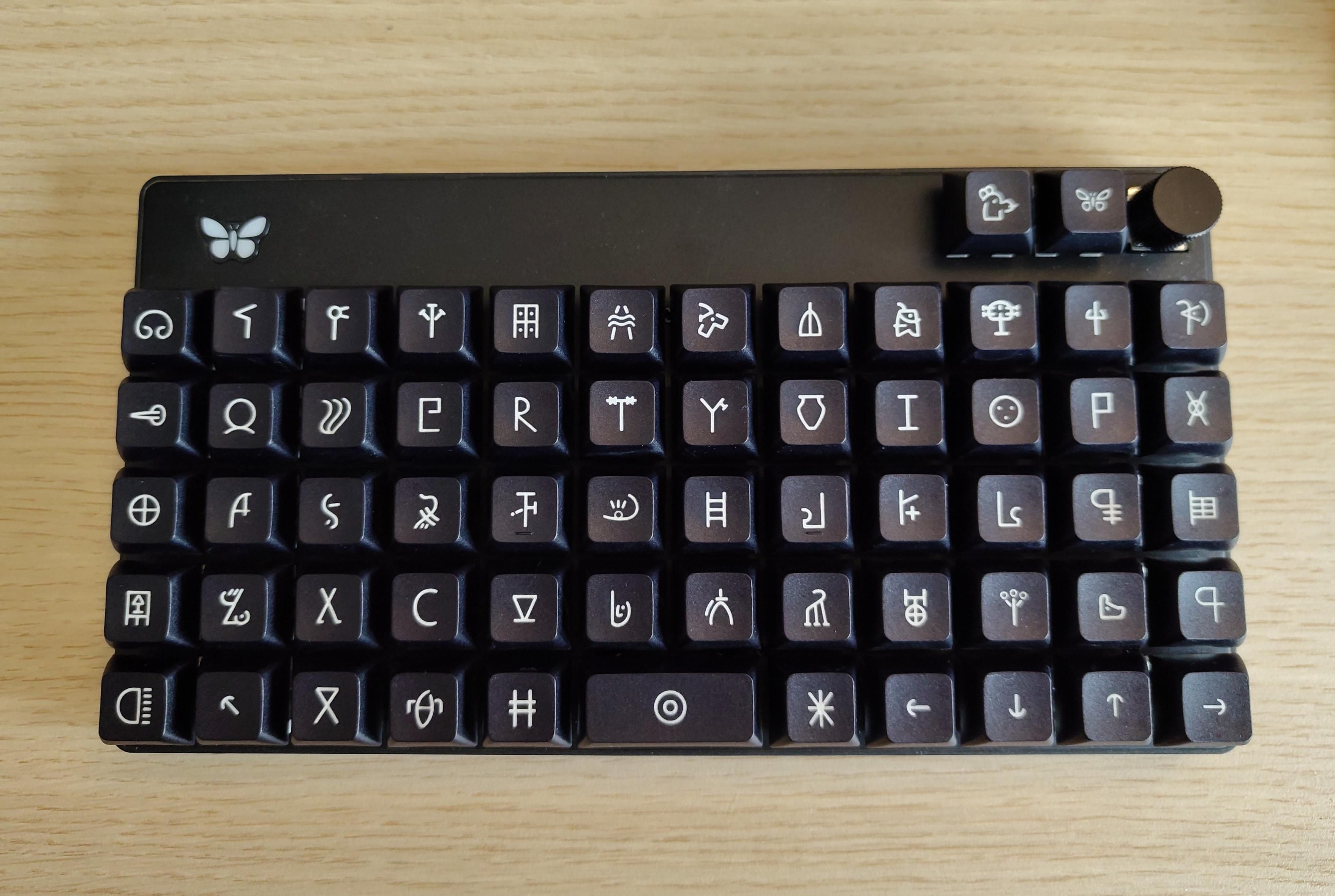 A black ortholinear keyboard in a 12x5 grid, with two buttons and a knob at the top right and a butterfly logo on the top left. The key caps are the undeciphered "Linear A" character set, a writing system used by the Minoans of Crete between 1800 BC and 1450 BC. Some characters bear a resemblance to modern Latin characters like C or R. Others look like animals or more abstract shapes