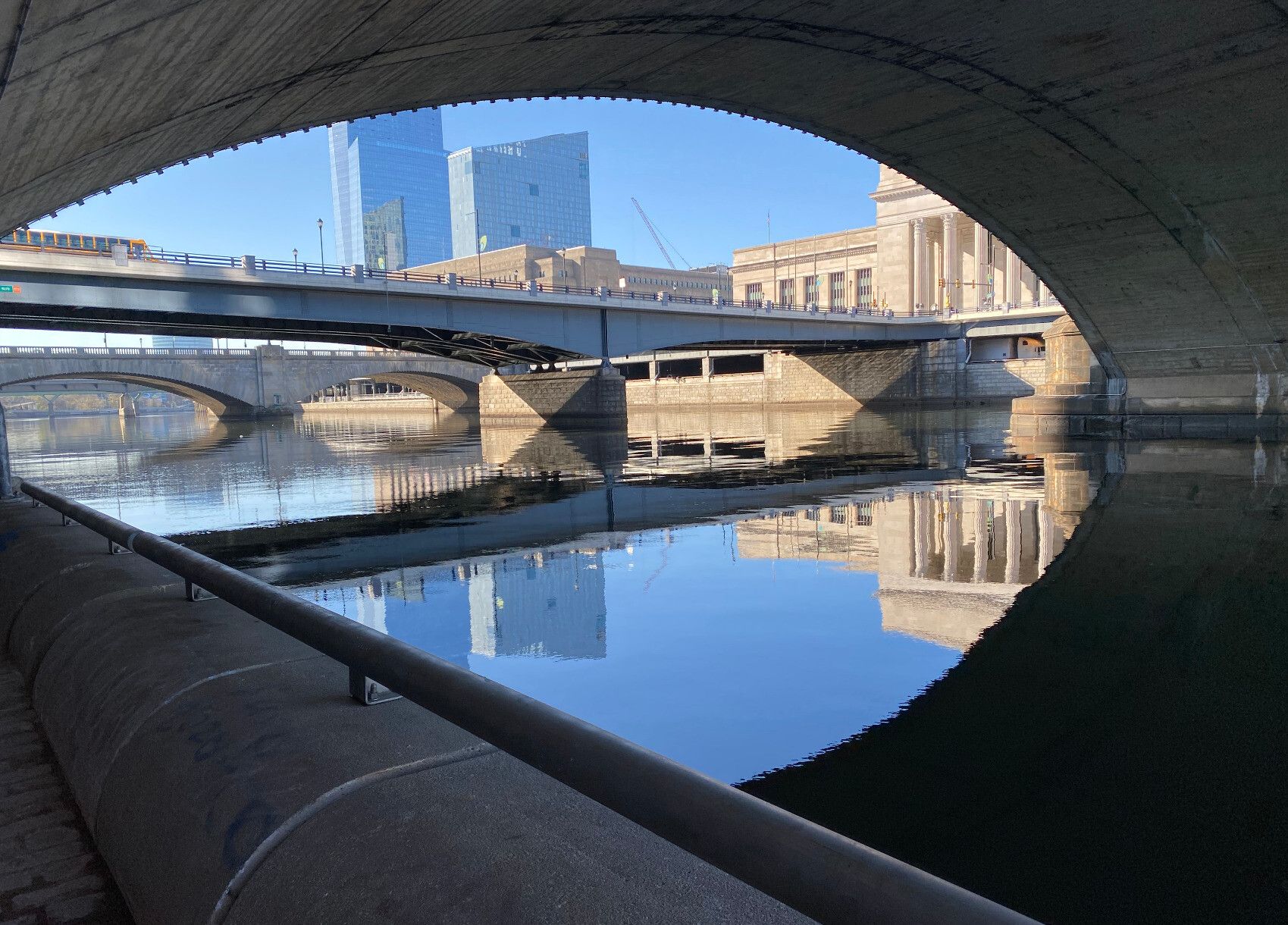 View of the Schuylkill River from the Schuylkill River Trail -- perspective is under one bridge (arching across the top of the image) and looking down river at several more.  The bridges appear in shades of tan stone, with some blue metal; the sky above and water below are blue; and there are buildings visible above the bridges -- two modern blue glass and one, to the side, with classical sandstone facade and columns.