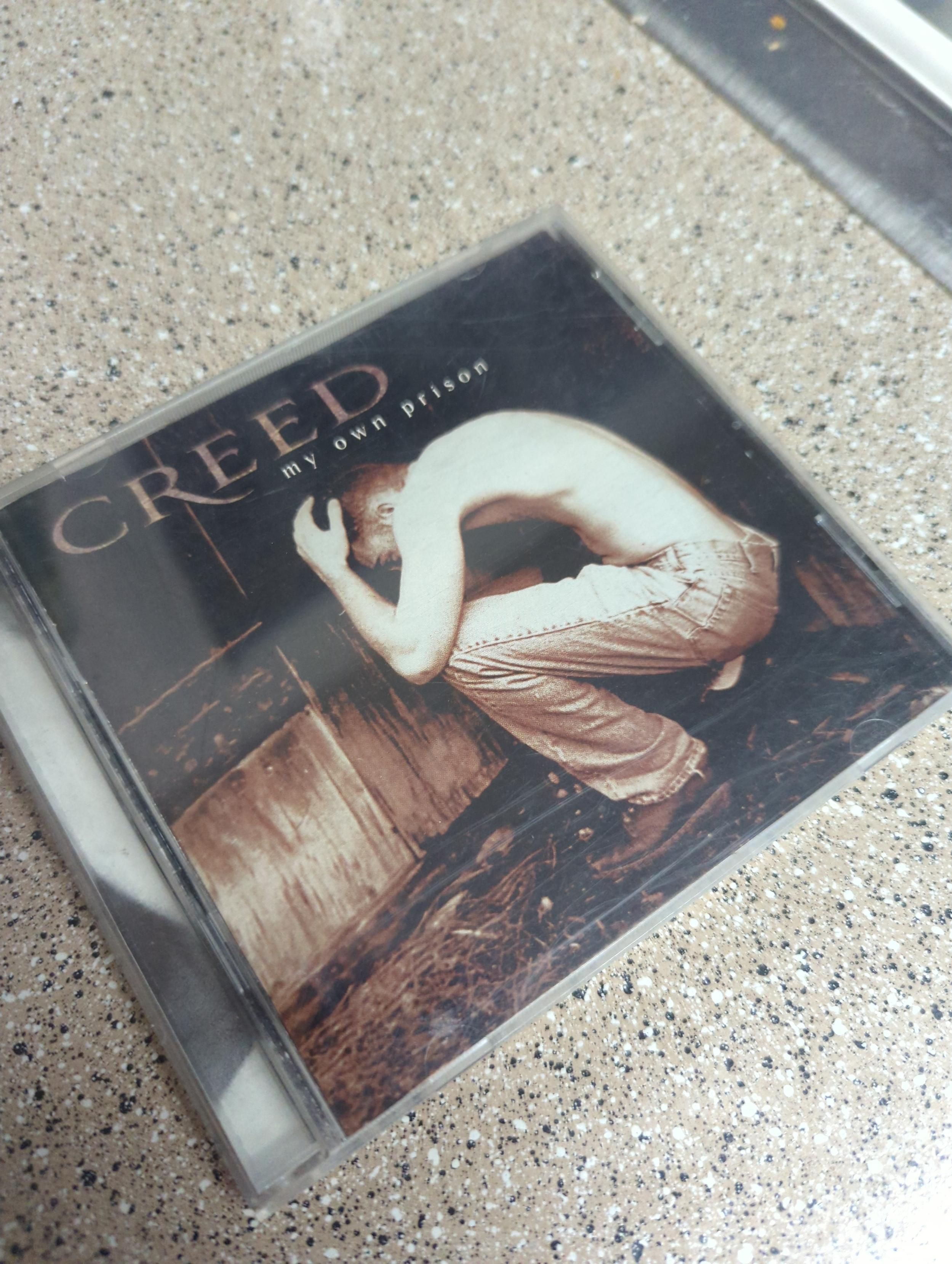 A CD case with a sepia photo of a shirtless man crouched in the fetal position. Above him, it says "CREED my own prison". The clear plastic case has subtle but noticeable wear. This CD has been places, clearly.