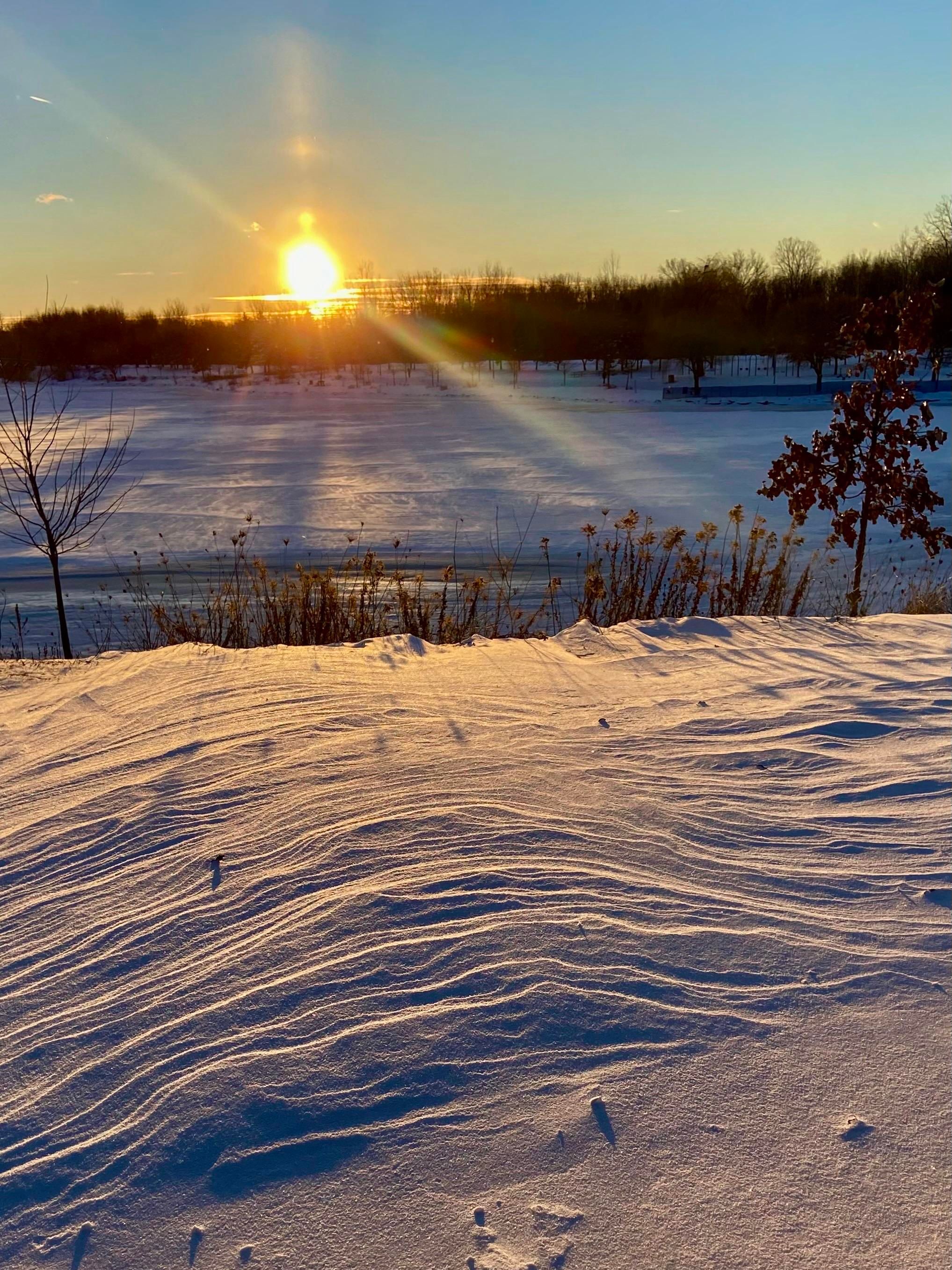 Wind rippled snow is in the foreground, a frozen pond surface beyond. The sun is setting, and it's sending out sunbeams (ok, fine, camera flares) up and to the left and down and to the right. Plus an apparent beacon going straight up. 