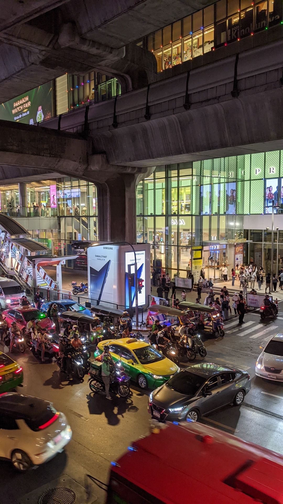 Outside Siam Paragon Mall at night. It's the central BTS SkyTrain transfer point, as well as the nexus of a chain of 3-4 malls. And a busy intersection. Two lines of the SkyTrain are near the top as cement overpasses. The colorful glass front of Paragon Mall featuring a Prada store is in the center. At the bottom is Sukhumvit Road, with a melee of cars, taxis, and motorbikes.