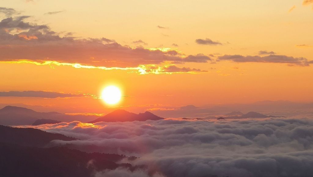 A golden sunrise rises above a sea of clouds, casting warm orange and pink hues across the sky and silhouetting distant mountain peaks. The sunlight breaks through scattered clouds, illuminating the landscape with a soft, ethereal glow.