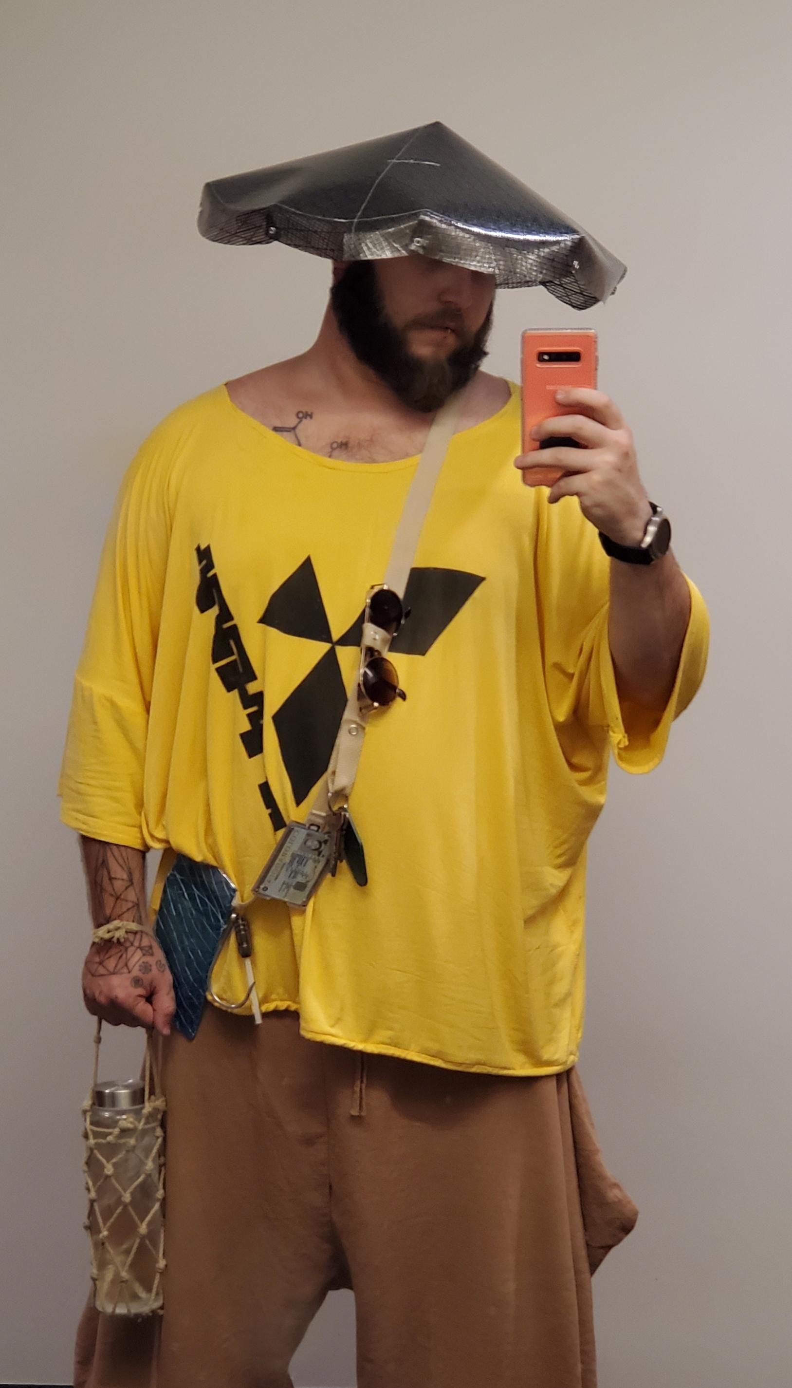 Selfie. Yellow, oversized t-shirt with a black logo. Wearing a bag made from nylon webbing and sail polymer and a conical hat also made from sail polymer. Holding a glass water bottle in a rope net. 