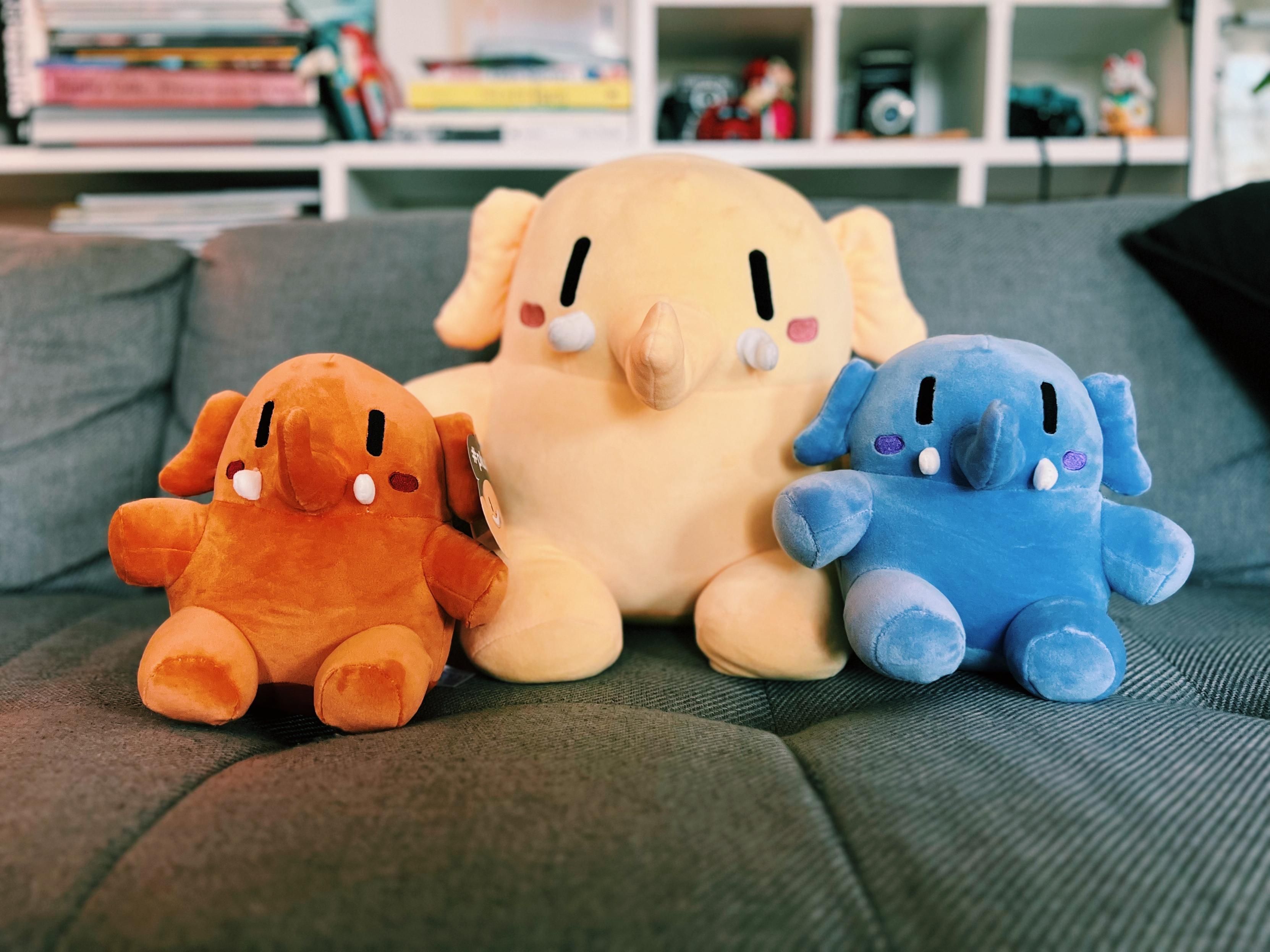 A photo showing three Mastodon plushies sitting on a couch. From left to right: a small orange one, in the center the original big beige/yellow one and on the left a blue one 