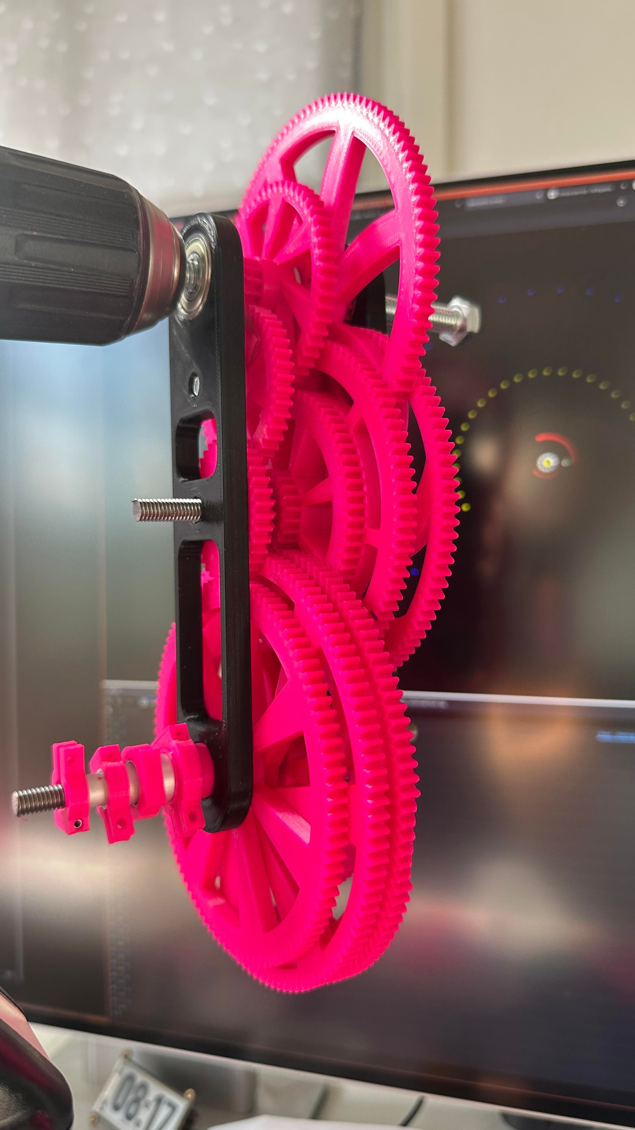 Complex geared assembly hooked to a drill chuck, held next to a screen showing orbits of planets. 