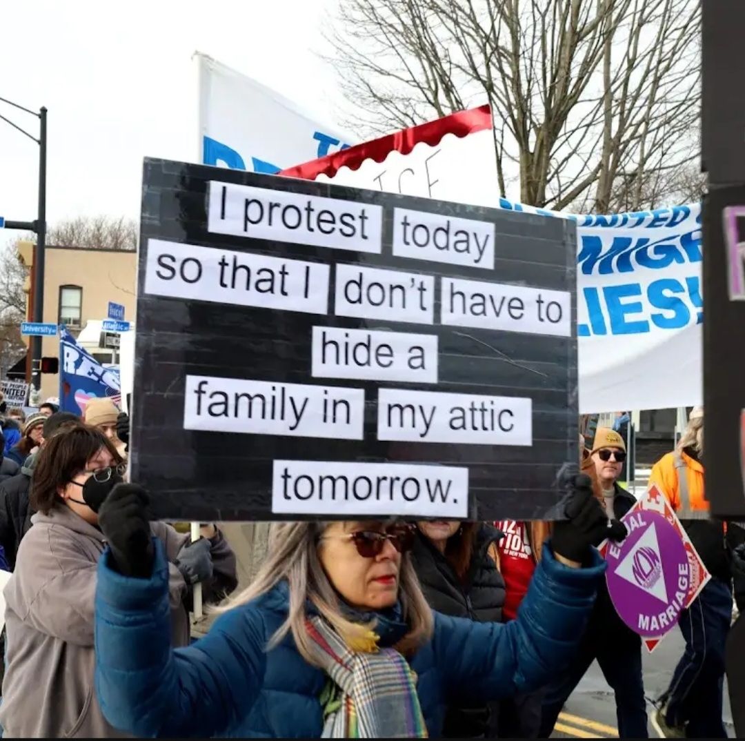 A woman  carrying a protest sign that reads: I protest today so that I don't have to hide a family in my attic tomorrow 