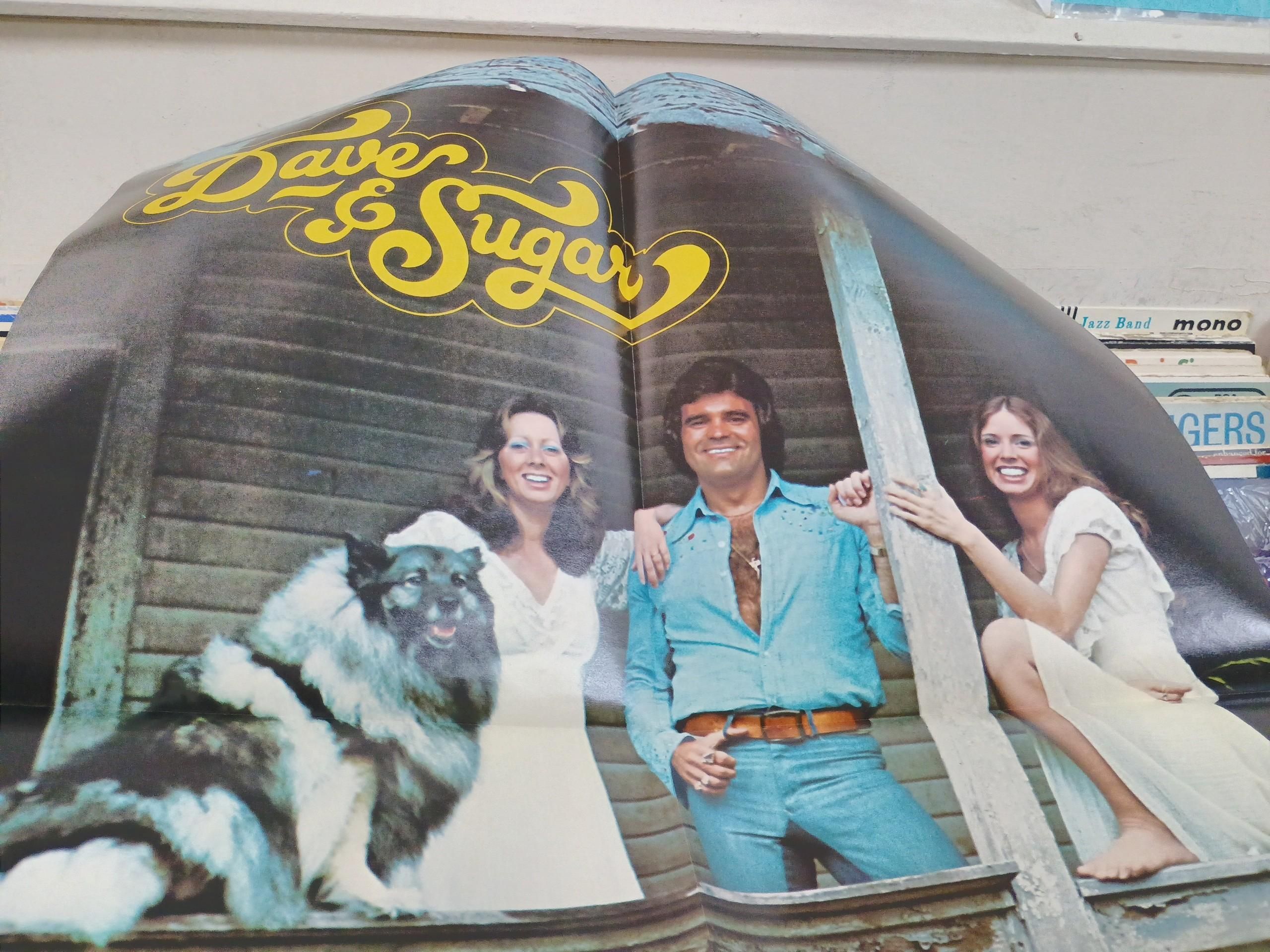 A large folded poster shows the same singing group of two gown -wearing ladies with old Dave and his shirt open half way revealing the wooly marmot that lives within.