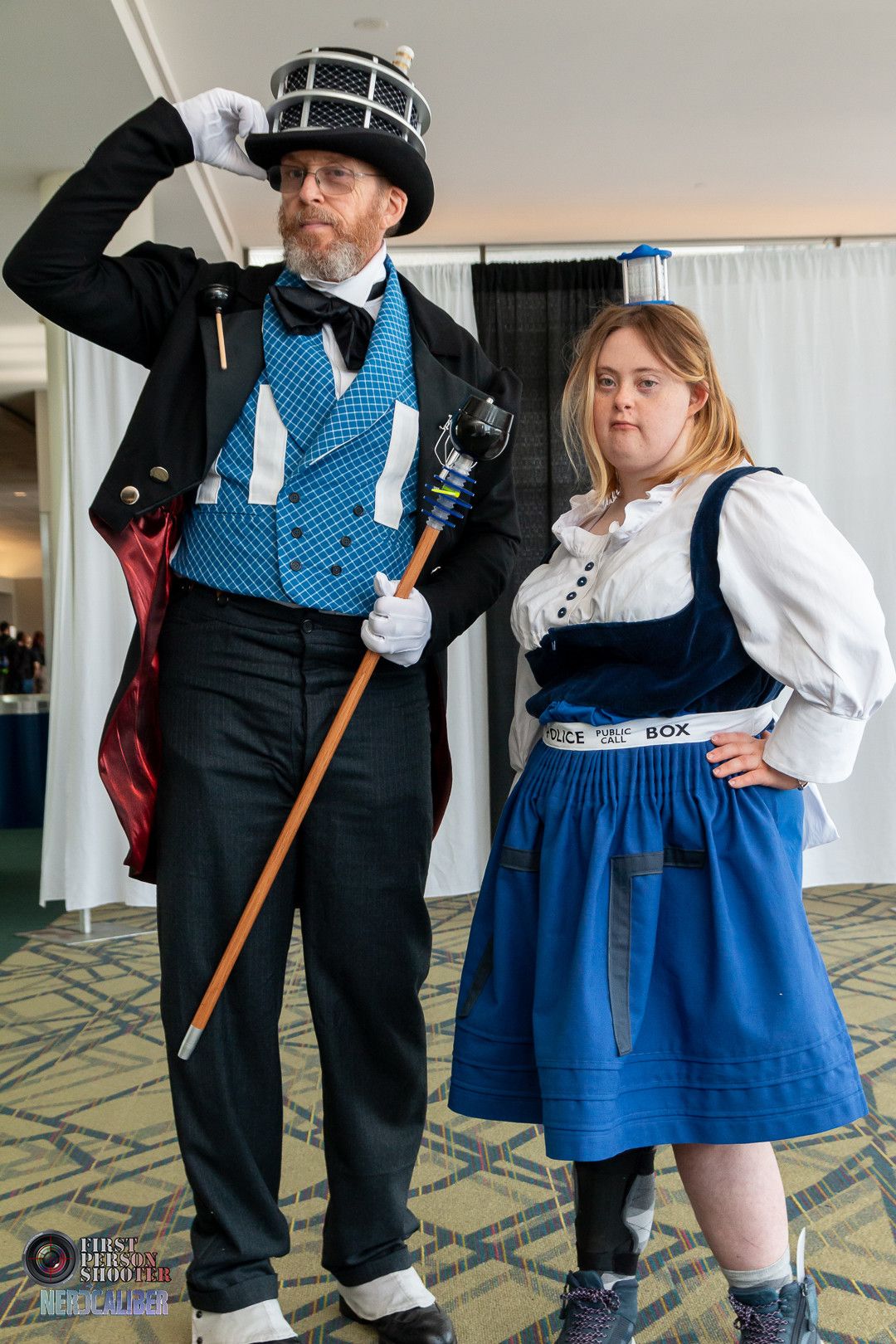 A man cosplaying as a Dapper Dalek wearing a full suit, tailcoat and top hat next to a young woman with Down syndrome cosplaying as a TARDIS