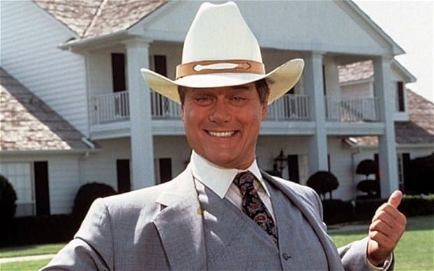 A suited and Stetson-hatted Larry Hagman as JR Ewing, grinning widely and thumbing at the Southfork Ranch house behind him. 