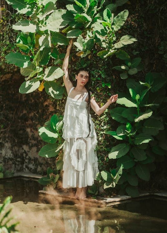 A fem figure dressed in floaty white layers and with one long braid of dark hair over their shoulder, standing on the edge of a pool against a wall, reaching up to the huge leaves of a plant growing overhead. They're standing in dappled sunlight.