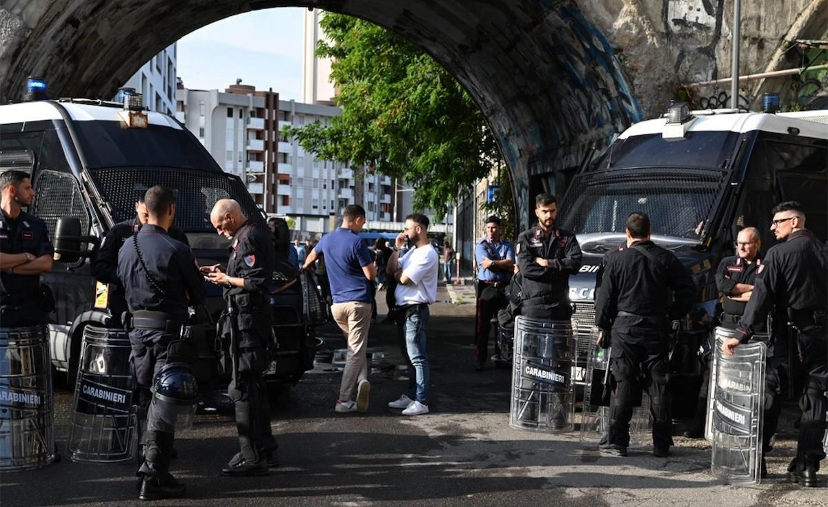 Squadra di agenti dei Carabinieri in assetto antisommossa appostata sotto un cavalcavia: una decina di uomini in divisa con elmetto e scudi anti-sommossa, due furgoni blu con il bollino dell’Arma, due uomini in abiti civili che parlano con gli agenti sul ciglio della strada; sullo sfondo edifici residenziali e l’intradosso grigio del ponte.