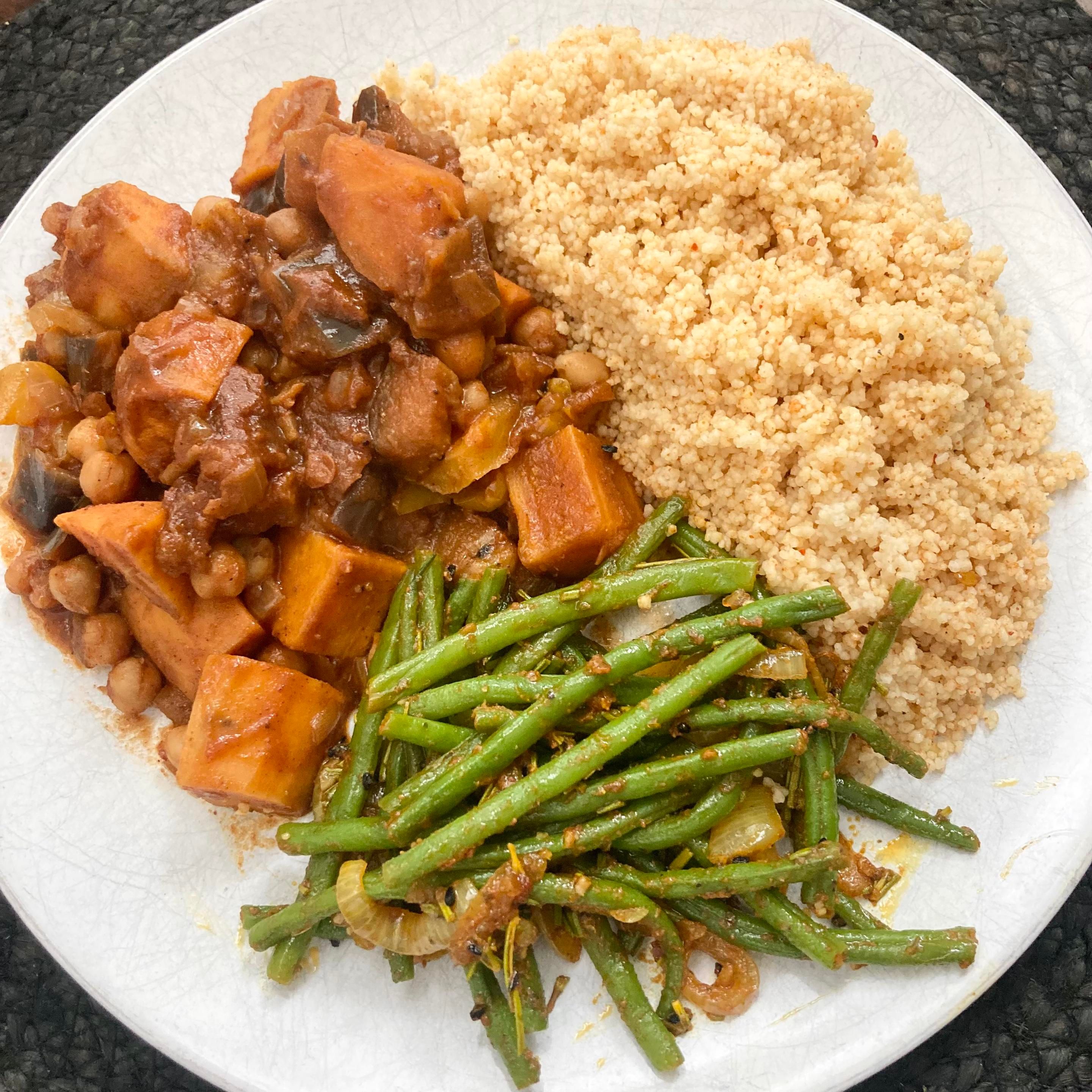 Overhead shot of a plate of food in three parts. To the top right a pile of cous cous flecked with harissa spice. Bottom right are green beans and onions that have a rosemary, cumin, turmeric and nigella seed blend. Top left is a tagine stew, deep orange with chunks of sweet potato, aubergine, chickpeas and dates.