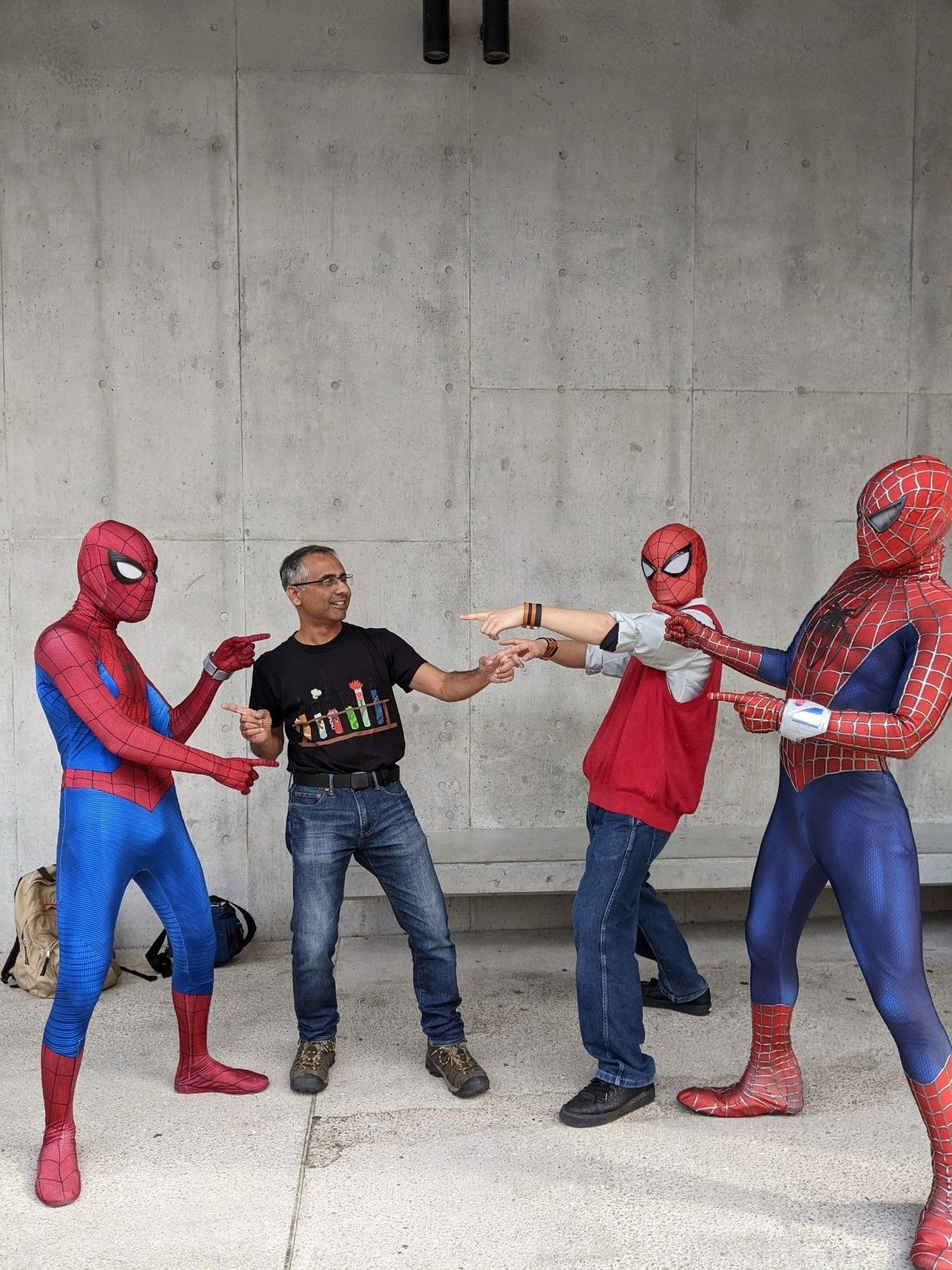 Three costumed spidermen and a person in a t-shirt pointing at each other