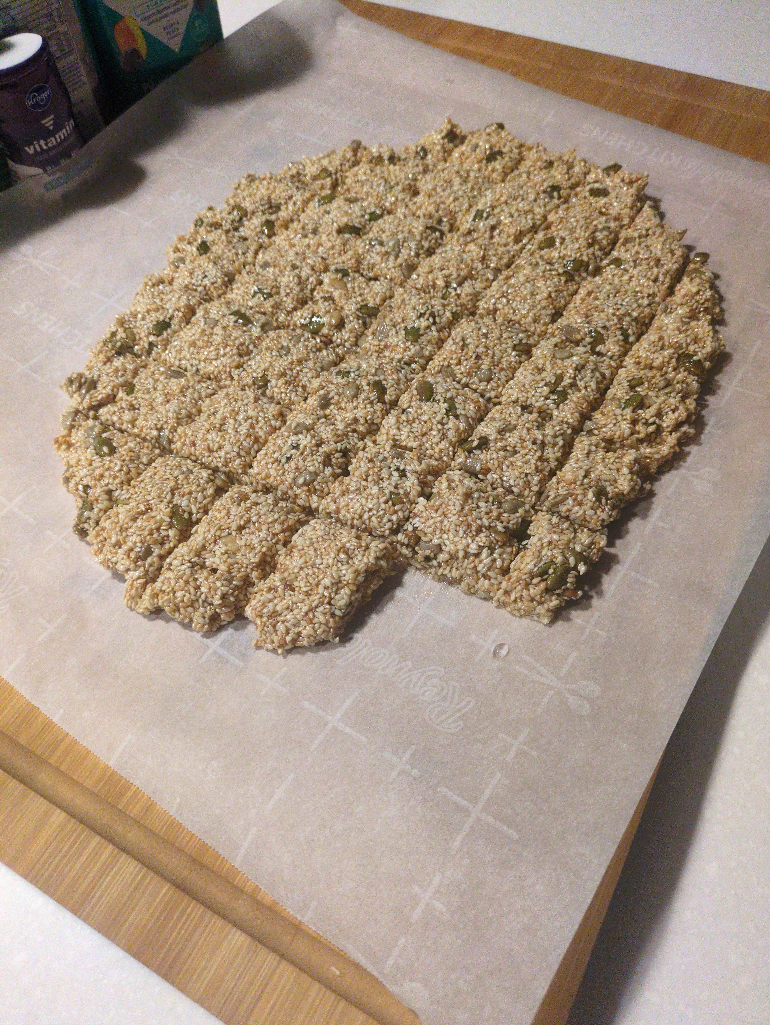 A cutting board with parchment paper, an oval of sesame seeds and honey, cut into segments.. 
