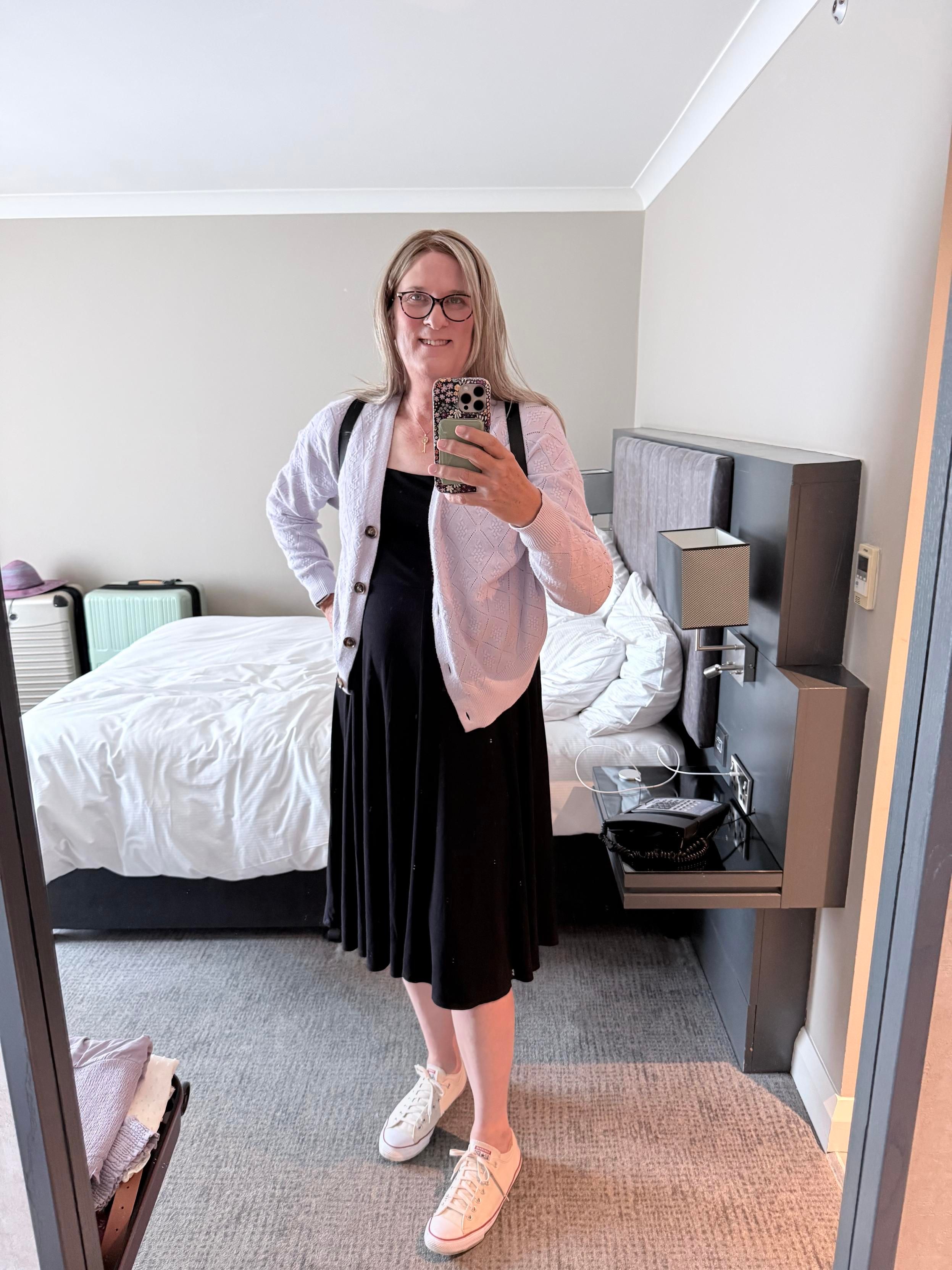 Photo of Sophie in front of a full-length mirror in her hotel room. She has shoulder-length blonde hair and is wearing black and pink glasses. She is wearing a black midi dress, a lavender cardigan, and cream coloured low-top Chucks. The black straps from her backpack purse are showing in the front. She is happy and ready to start the day!