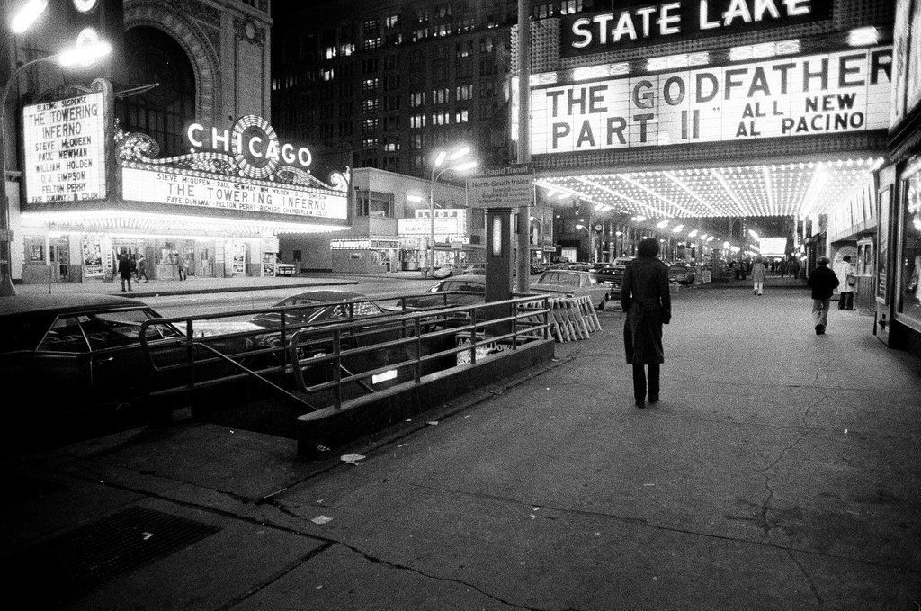 1975 photo of State Street, Chicago, at night, main subjects being the State Lake and Chicago theatres, both cinemas at the time, with one showing The Godafather Part II with Al Pacino and the other showing The Towering Inferno. Shadowy figures walk under the marquee lights, a somewhat "noir" image.