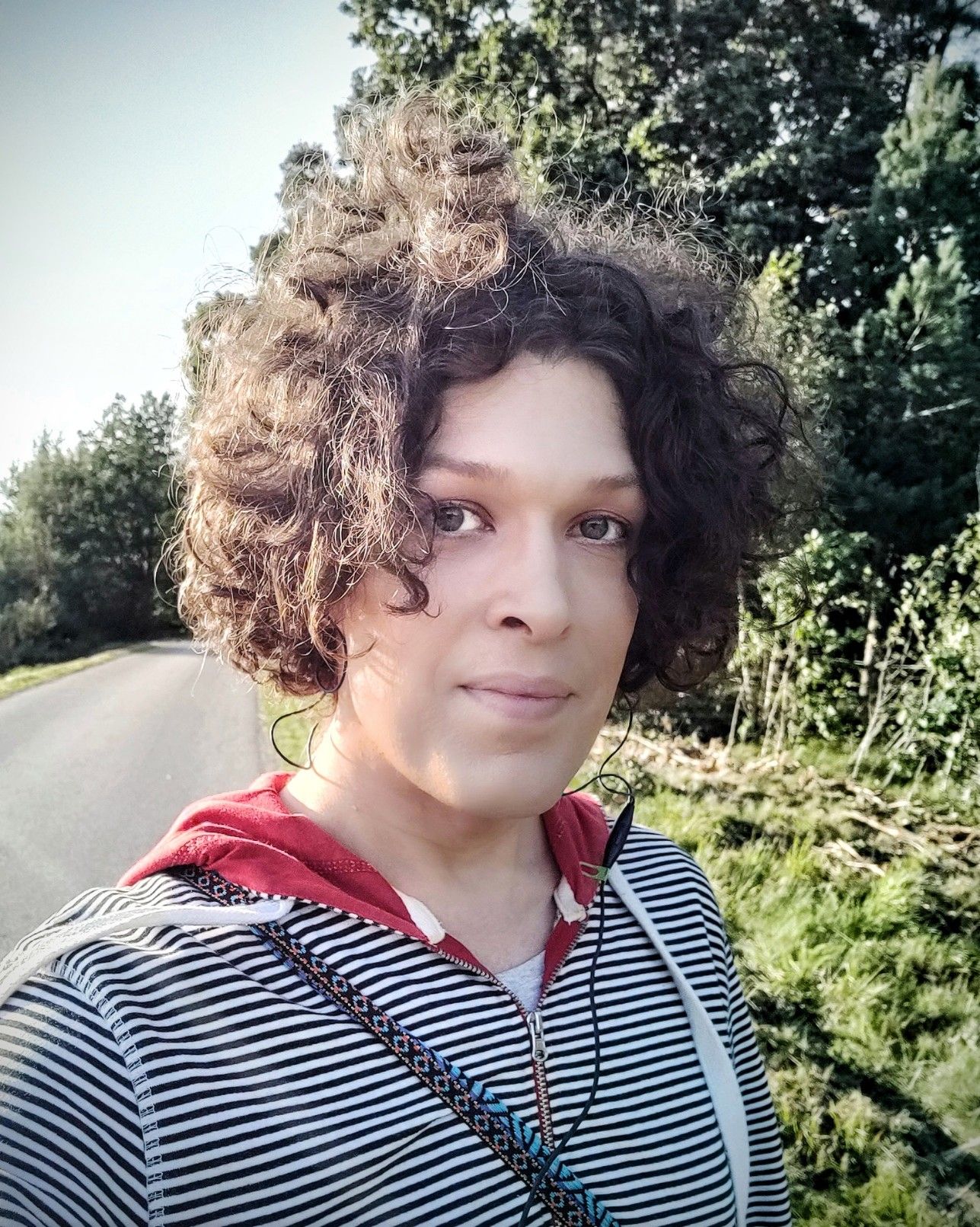 Selfie of me walking on a sunny road with leafy trees behind me. I'm wearing a black-and-white striped hoodie with red inside the hood.
