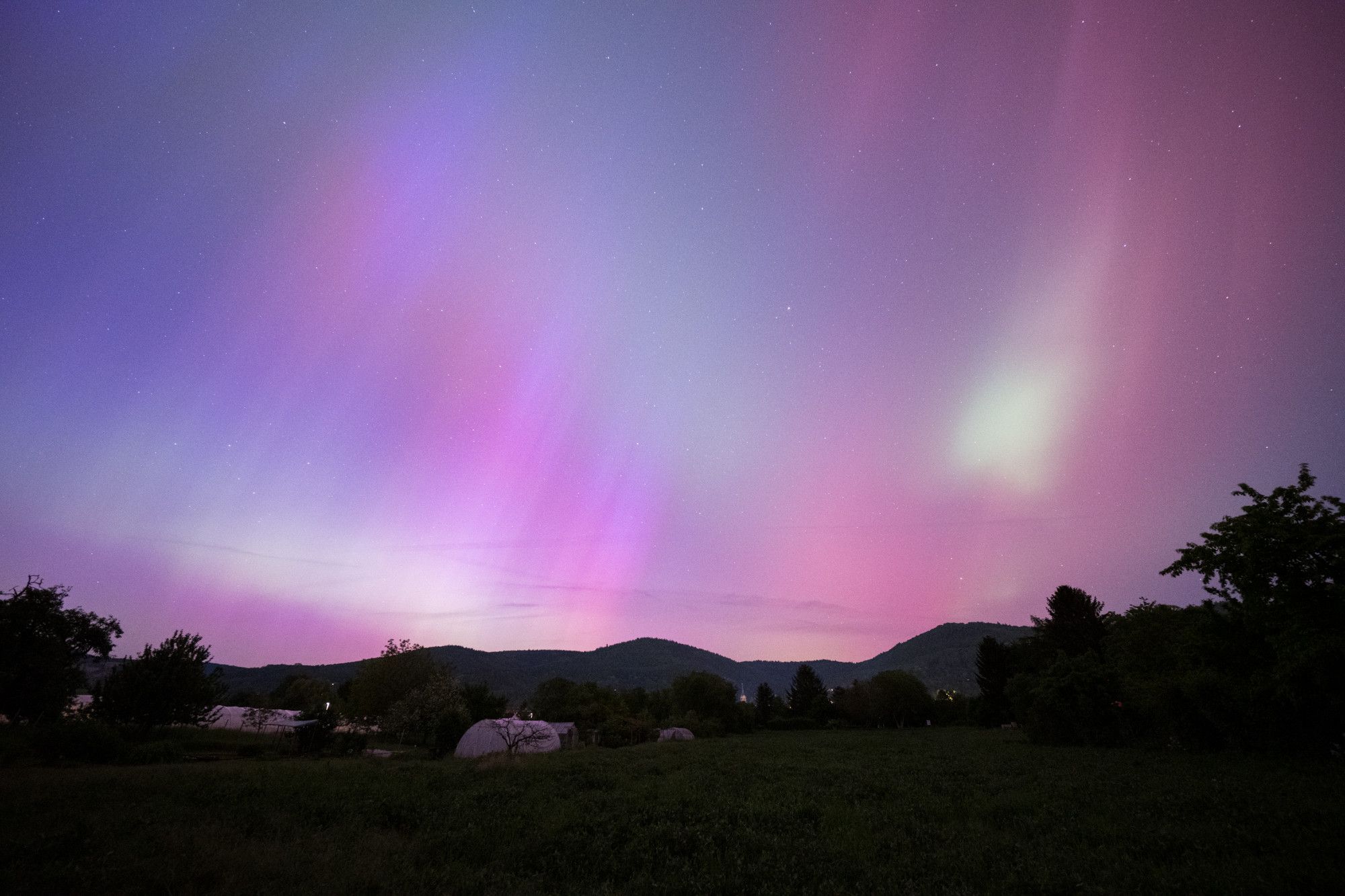 A field at night with a few medium-height mountains in the background (Odenwald near Heidelberg). The sky is illuminated by the Northern Lights, which appear as gentle veils and vary in color from violet-red to green-yellowish.  