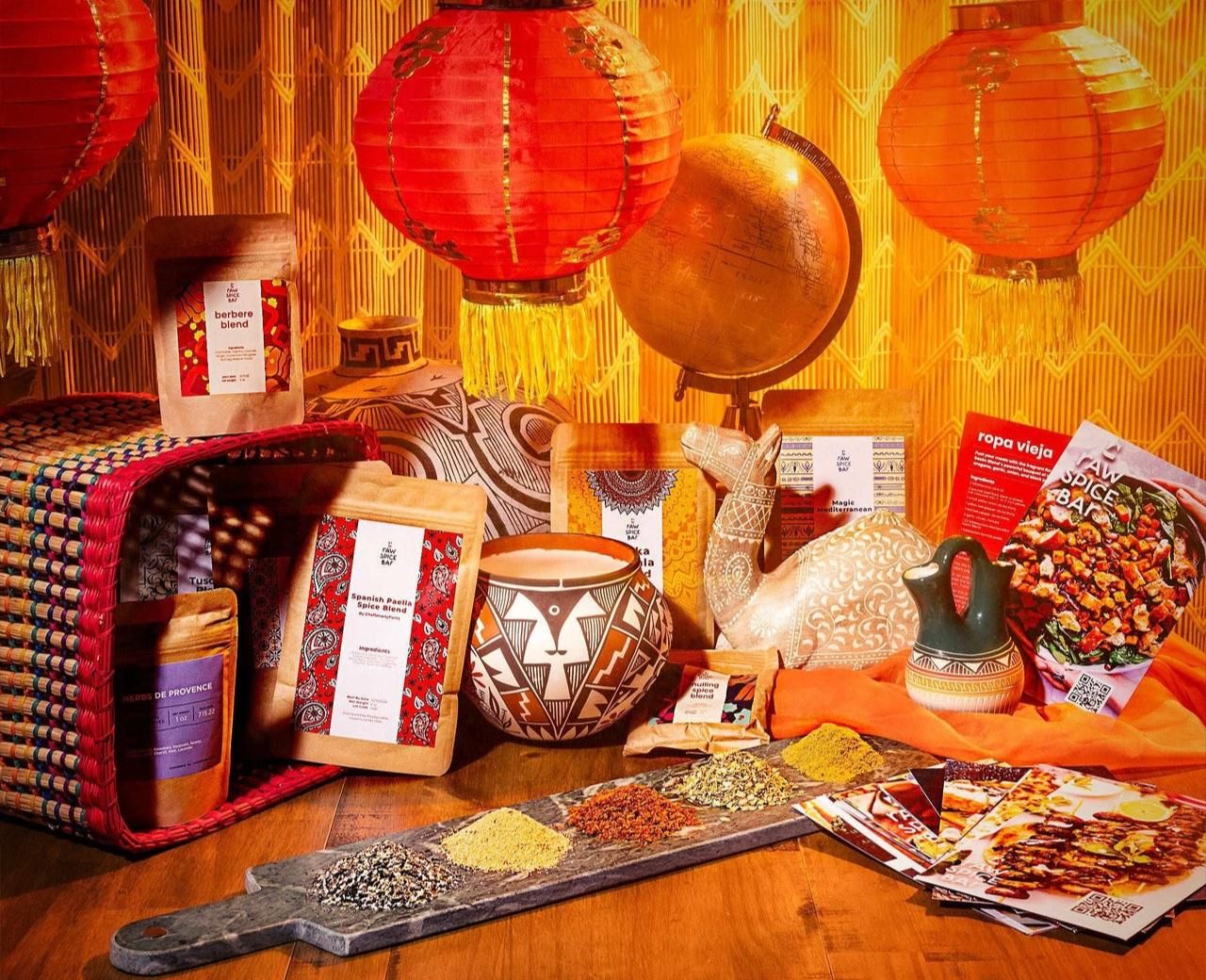 A vibrant, warm-toned still life featuring various global spice blends from Raw Spice Bar. Several brown paper spice pouches, including "Berbere Blend," "Spanish Paella," and "Herbs de Provence," are arranged around a woven basket, decorative pottery, and a small camel figurine. In the background, three glowing red paper lanterns hang above a vintage globe. In the foreground, small mounds of colorful ground spices are displayed on a long slate serving board alongside recipe cards.
