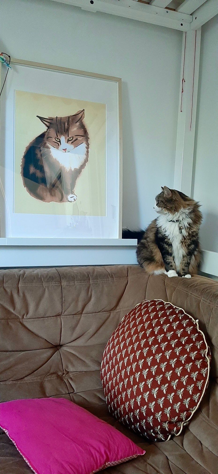 Chatte assise sur le dossier d’un canapé marron devant un portrait d’elle plus que grandeur nature que j’ai dessiné. Elle a la tête tournée vers son portrait, genre, c’est quoi ce truc ?