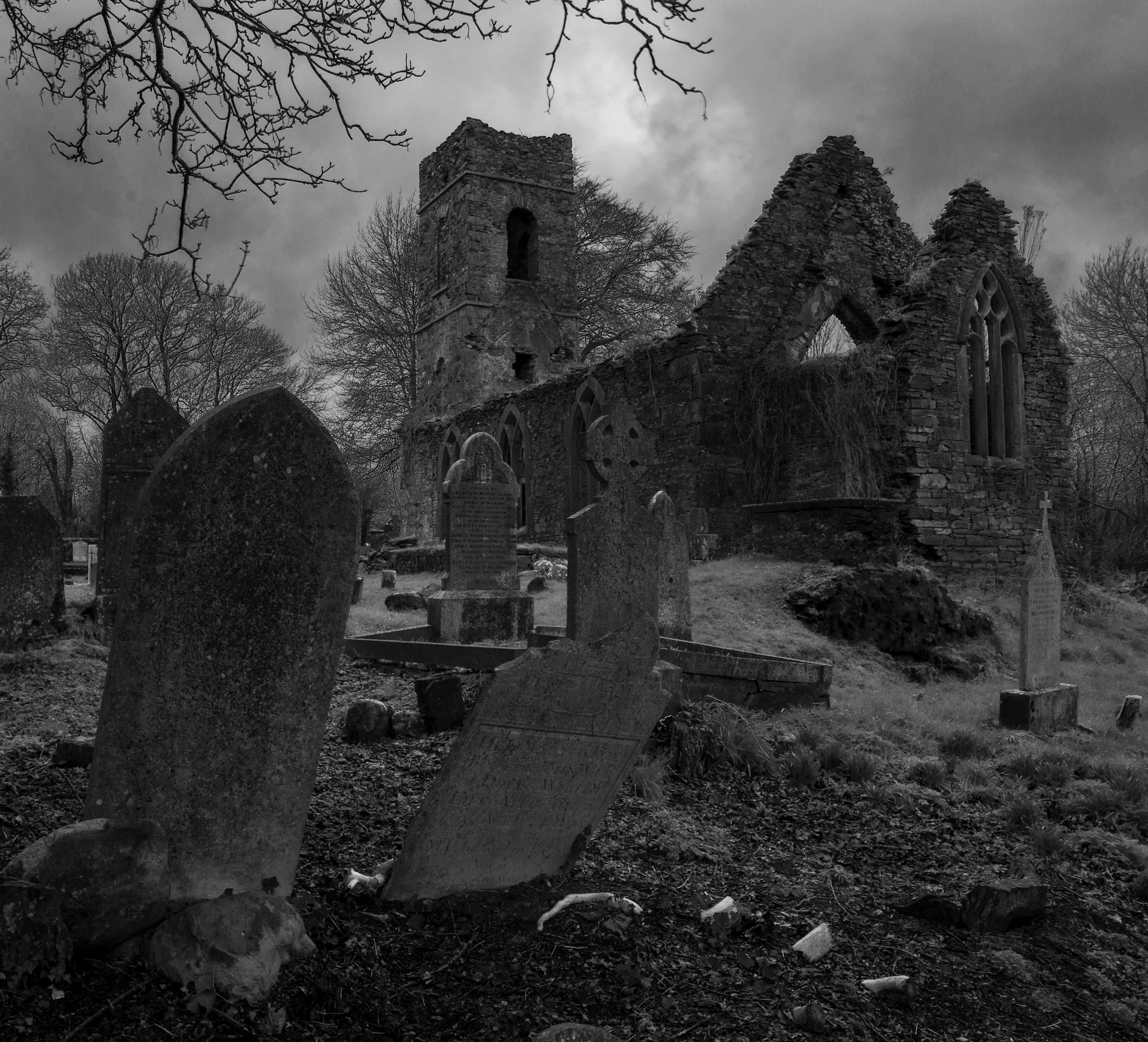 A black and white photo of an old ruined church, with gravestones in the foreground, some of which are tilted over.
The sky behind the church looks dark and forbidding and there are leafless tree branches in the top of the image.