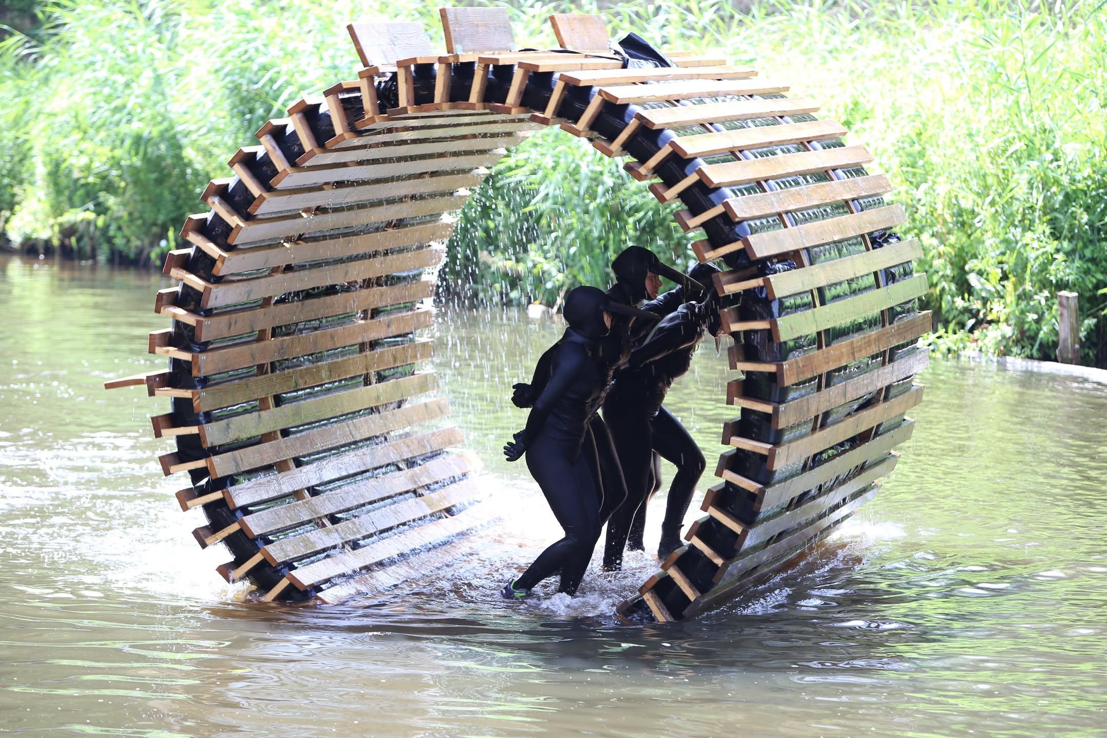 three people in skintight black outfits with beaks on their heads inside of a rotating cylinder that they are propelling by walking it across the water