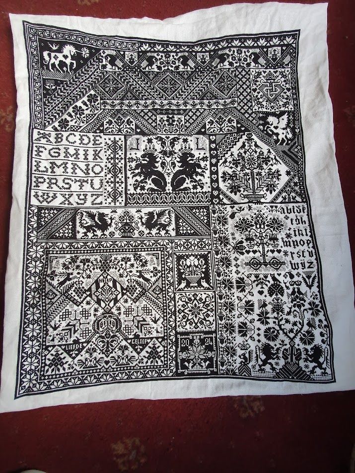 A large cross stitch project. 
Black stitching on white fabric. There is a solid black line border. The pattern is a sampler so includes the alphabet, animals, random geometric patterns. 