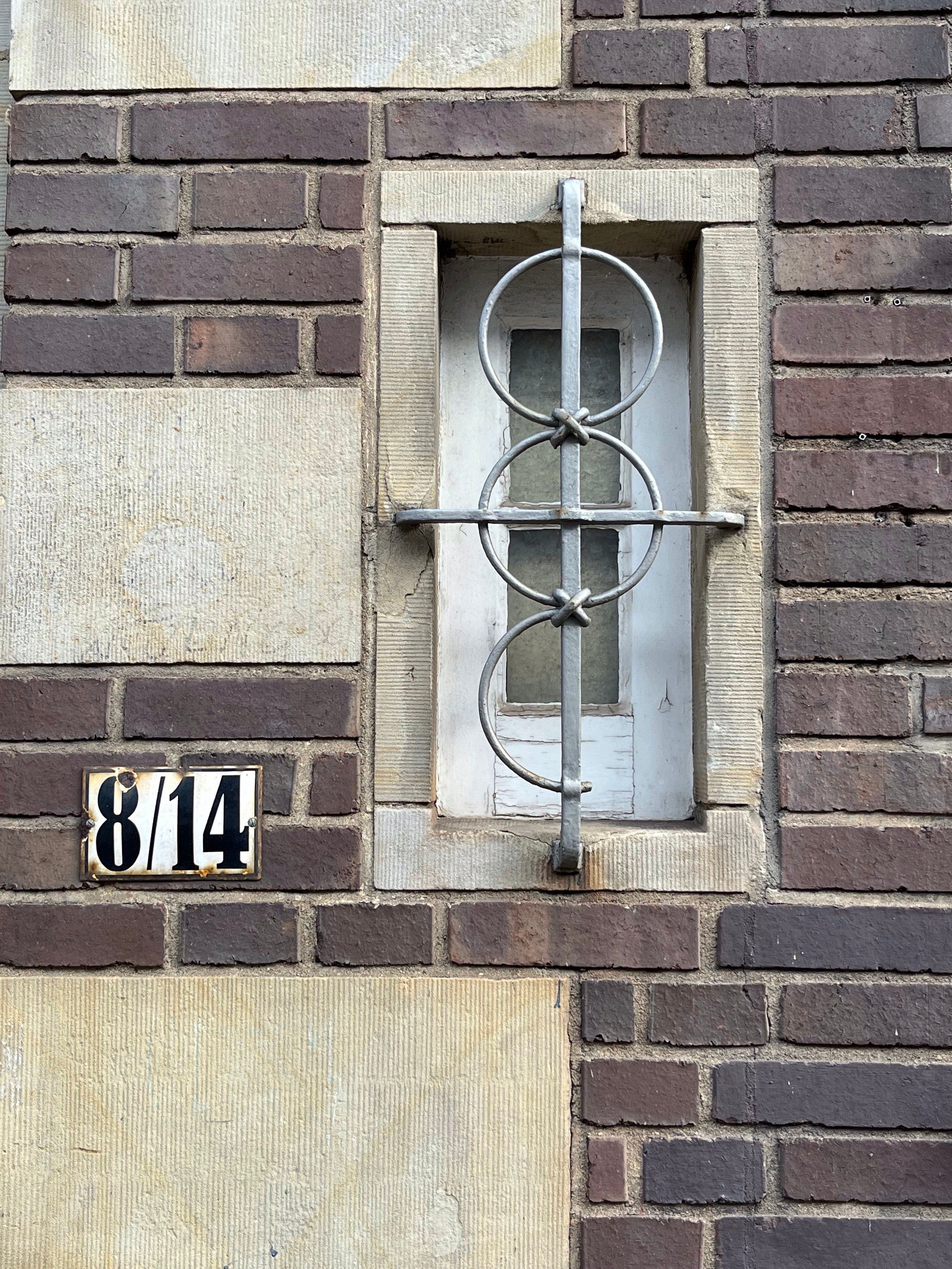 A close-up view of a patterned brick wall with a small window secured by a decorative metal grid. A number plaque reading "8/14" is positioned beneath the window.