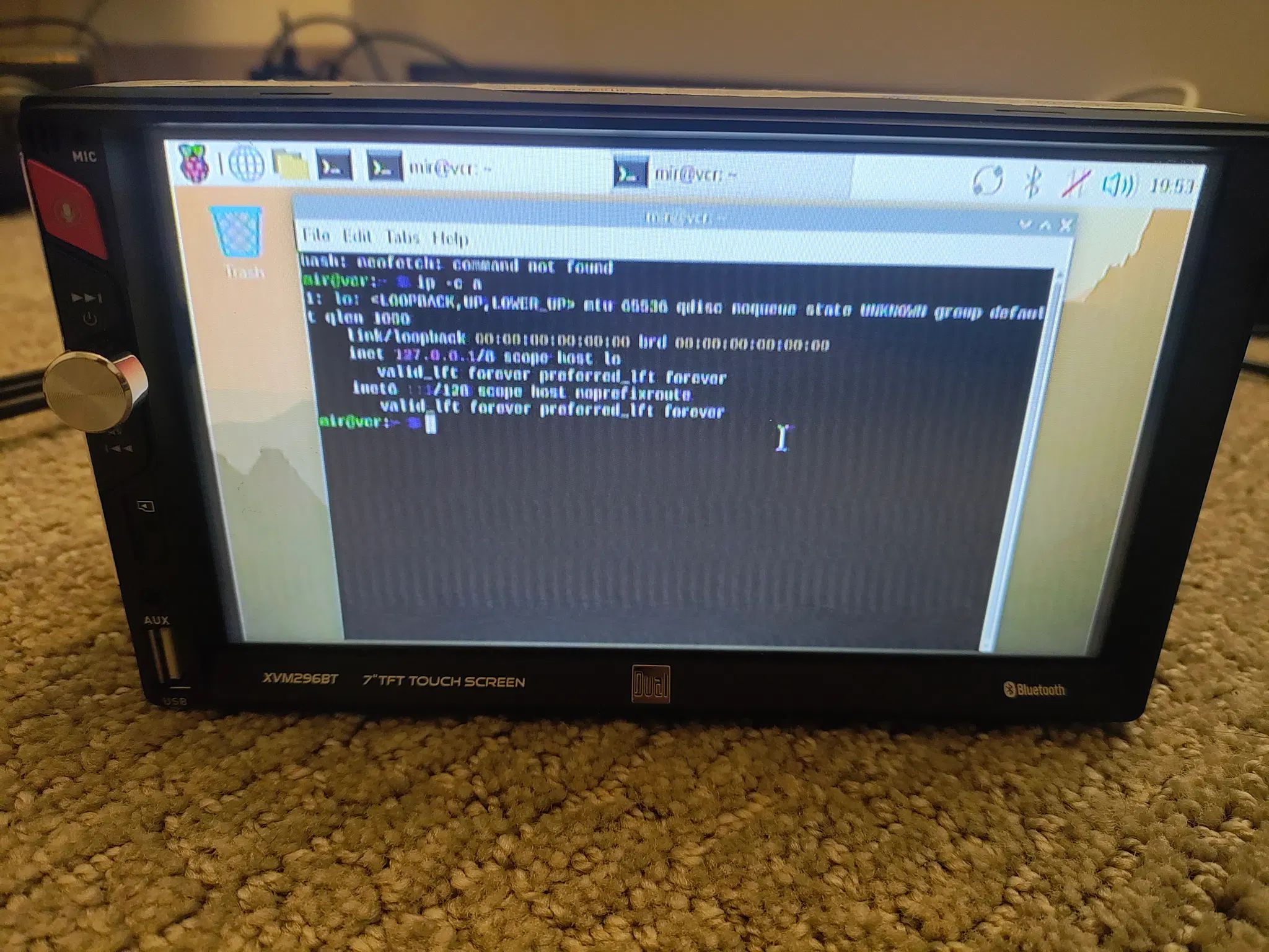 touch screen car stereo sitting on a carpet floor with an indistinct mess of wires in the background. it's a Dual-brand XVM296BT advertising a 7in touchscreen and Bluetooth. on the left bezel edge there are forward-backward buttons, a red mic button, a silver dial, and microsd, aux, and USB inputs.

the screen itself is showing
raspberry pi os with a terminal open. the user is mir and the hostname is vcr. the terminal shows a failed attempt to run neofetch, and the results of ip -c a (no network besides loopback, despite this being a pi zero W) 