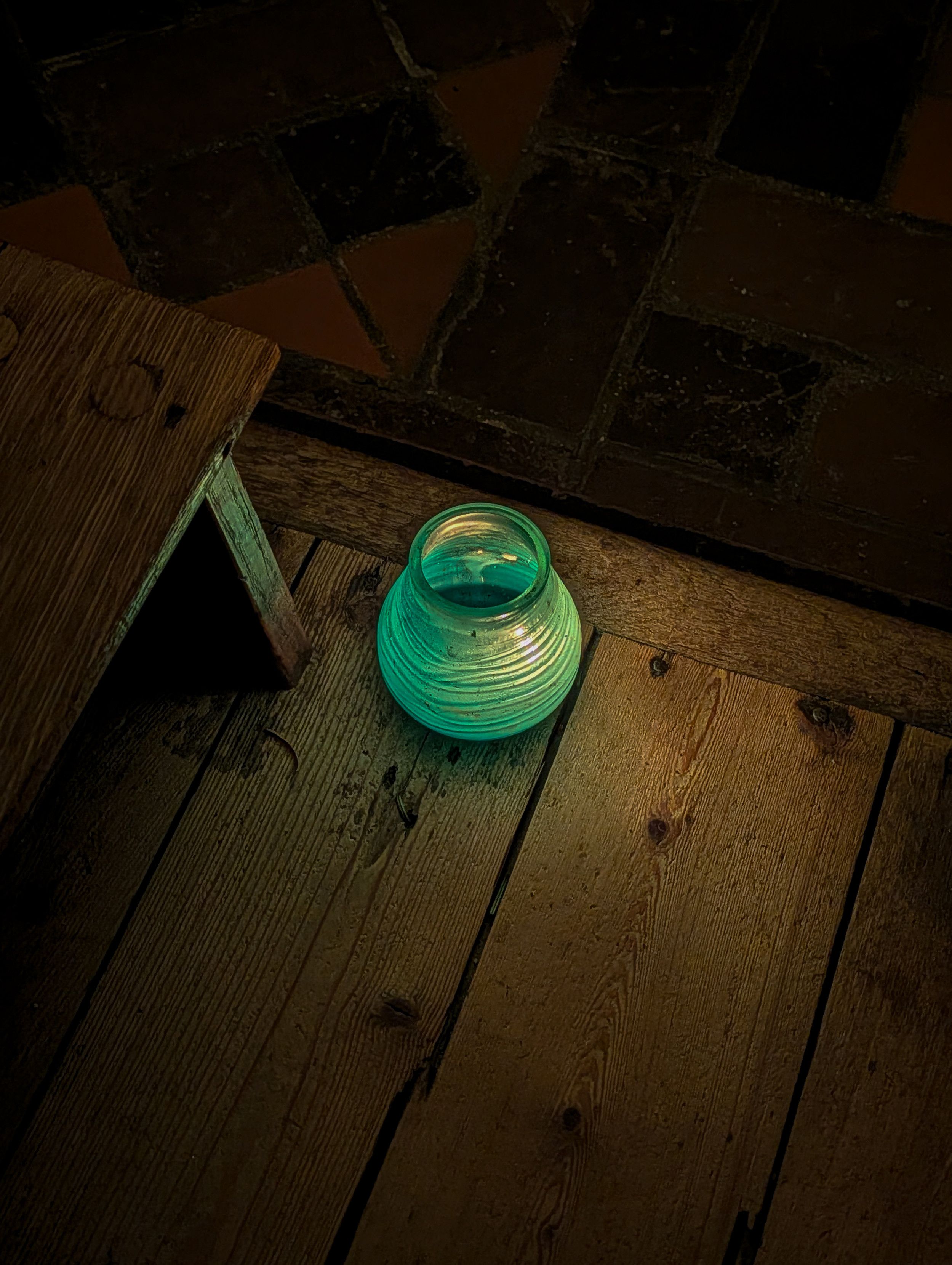A small, rounded green glass jar with a tea light inside. The lantern sits on weathered wooden floorboards. The wood grain is prominent, and the teal glow casts long, warm shadows and a colored highlight across the planks. 

To the left, a portion of a simple wooden church stool is visible. Behind the wooden platform is a transition to a darker tiled or stone floor.